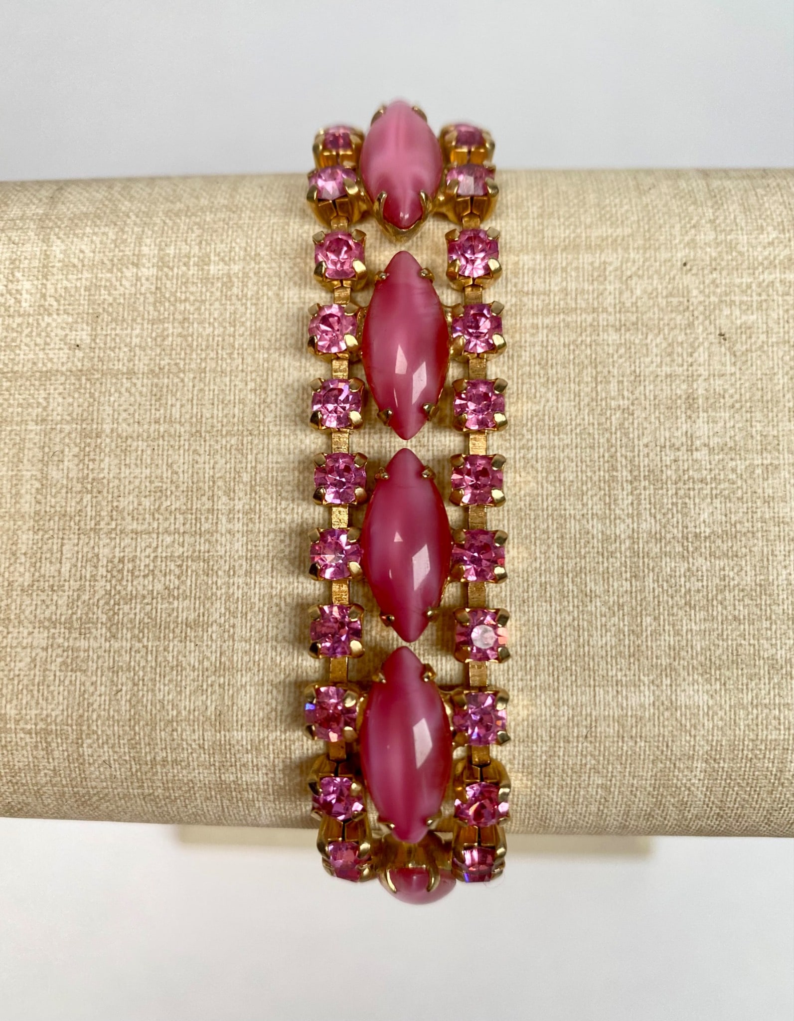 Vintage Gold Tone Bracelet with Pink Cabochons and Rhinestones: A gold tone bracelet featuring pink marquise shaped cabochons and pink rhinestones arranged in three rows. The bracelet is secured with a fold-over clasp and has an articulated design for flexibility.