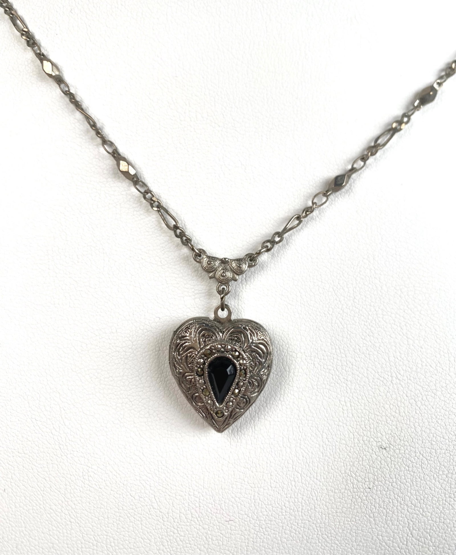 Vintage Heart Locket Pendant With Intricate Chain: Silver tone necklace featuring a heart shaped locket pendant with intricate decorative details and a black teardrop shaped stone at its center. The locket opens to reveal two compartments with a flora