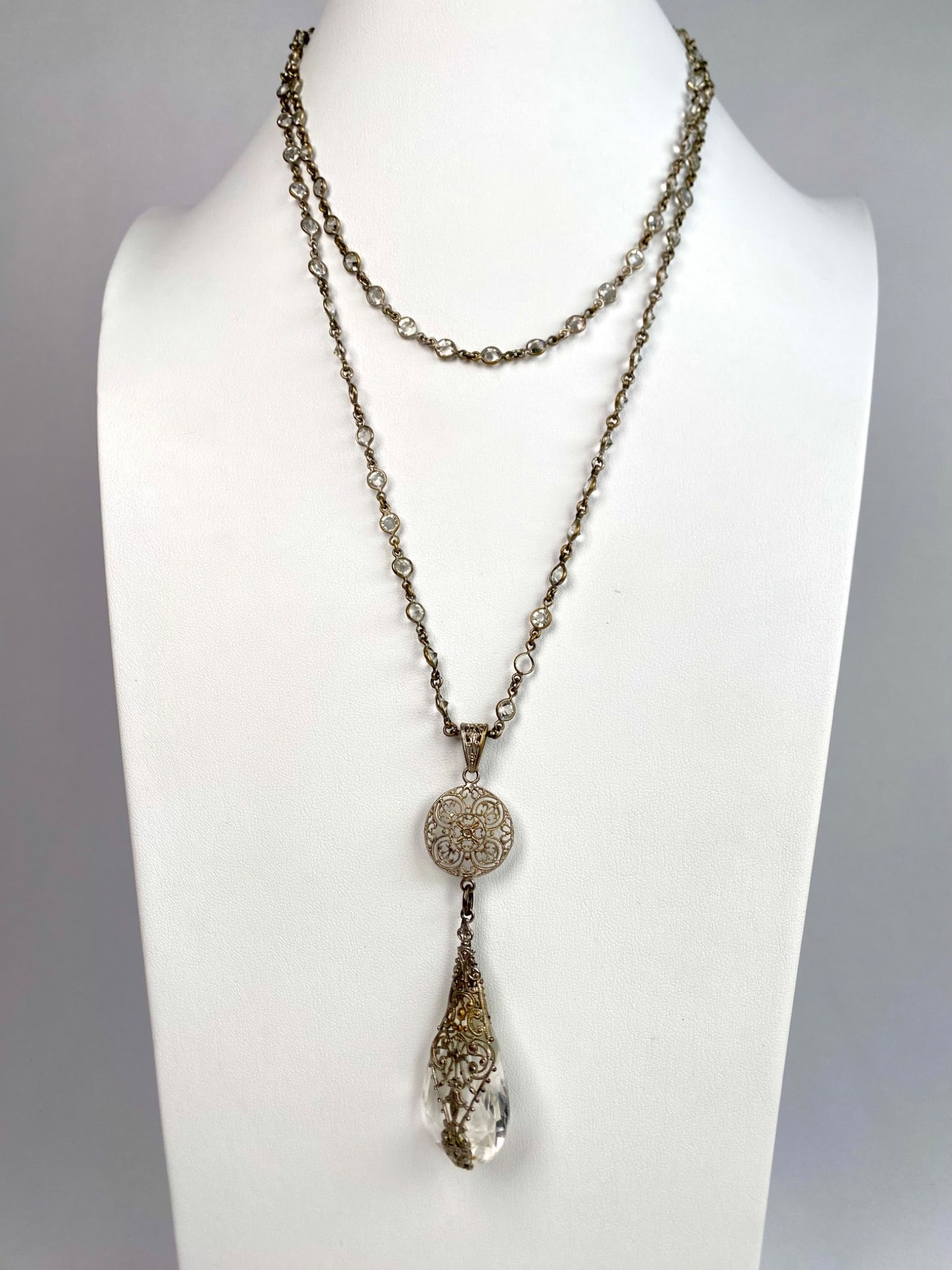 Art Deco Crystal Drop Pendant Necklace: A silver tone necklace featuring a long strand chain adorned with bezel set crystals. The centerpiece includes an intricately designed circular medallion with filigree detailing, suspending a large fa