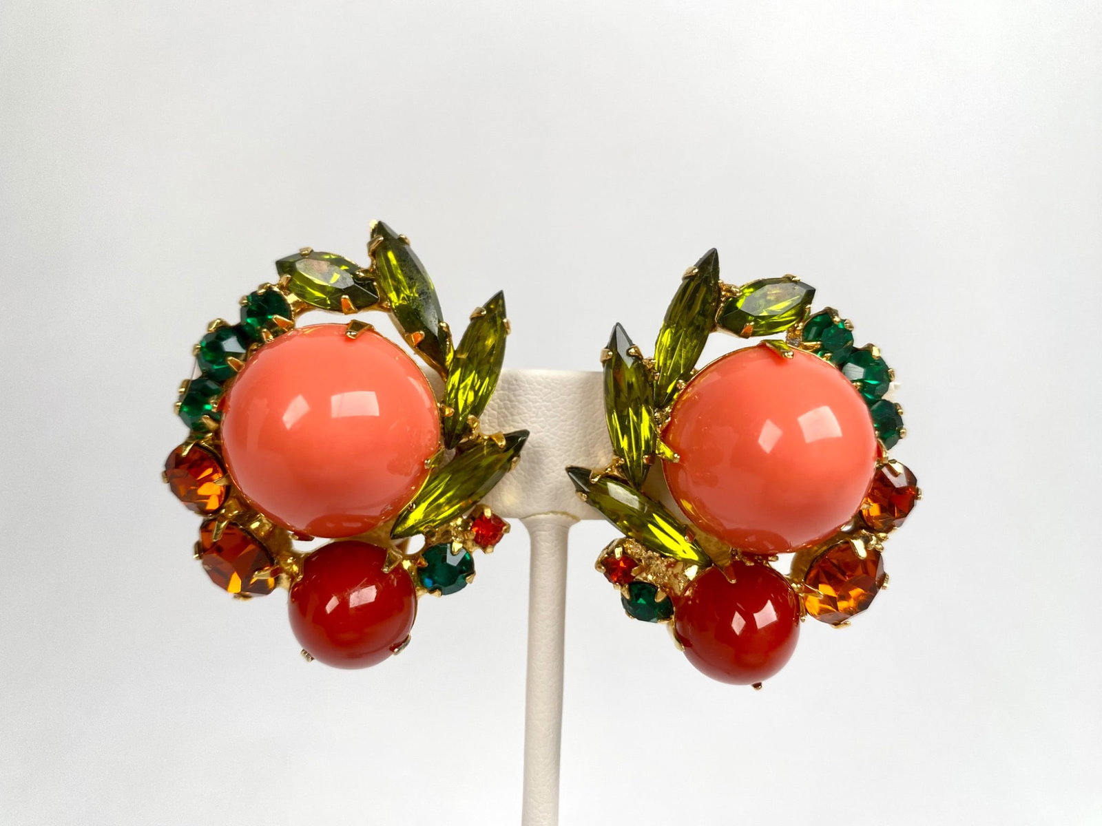 Pair of Gold Tone Clip On Earrings with Large Coral Colored and Red Cabochons: A pair of clip on earrings crafted from gold tone metal and adorned with large coral colored and red cabochons. Each earring features additional green, orange, and yellow rhinestones arranged in a flo