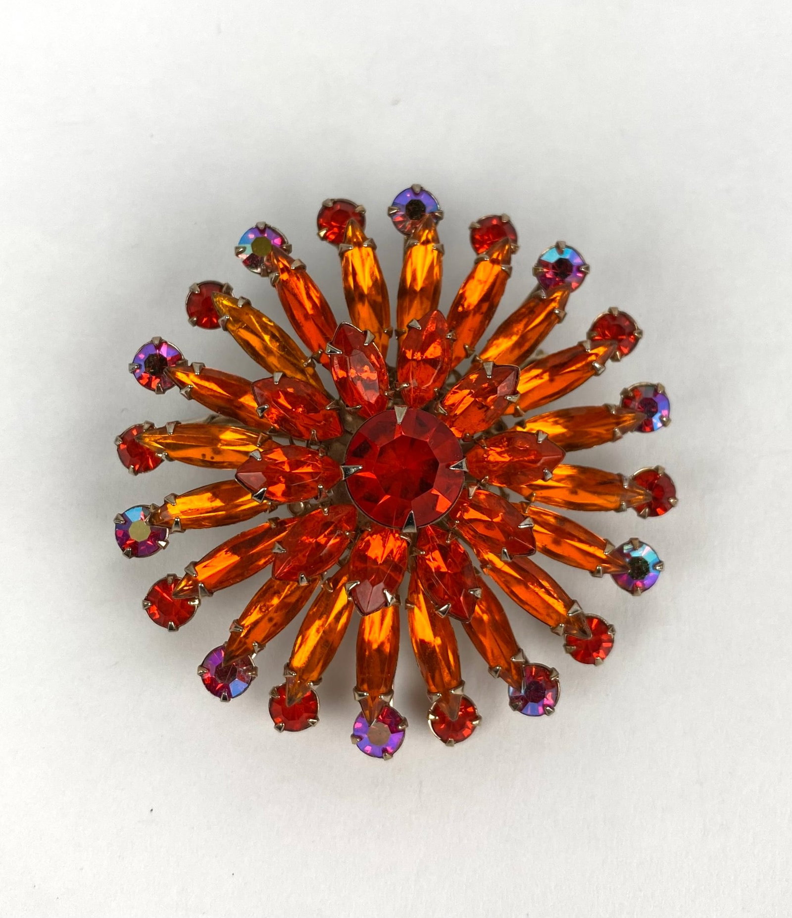Gold Toned Starburst Brooch With Orange And Red Glass Stones: Gold toned starburst shaped brooch adorned with faceted orange and red glass stones, with additional chromatic accents. The reverse features an intricate metal design with a locking pin mechanism for 