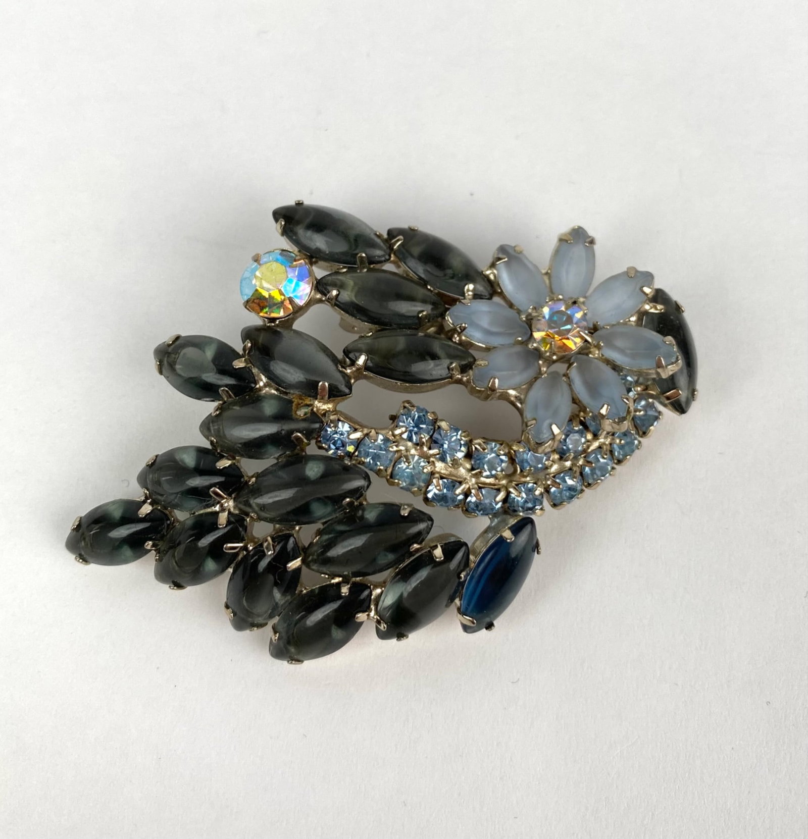 Art Deco Floral Black and Blue Rhinestone Brooch: Floral rhinestone brooch featuring various marquise, round, and navette shaped stones in hues of blue, gray, and aurora borealis mounted in a silver tone metal setting. The design includes a flower mo
