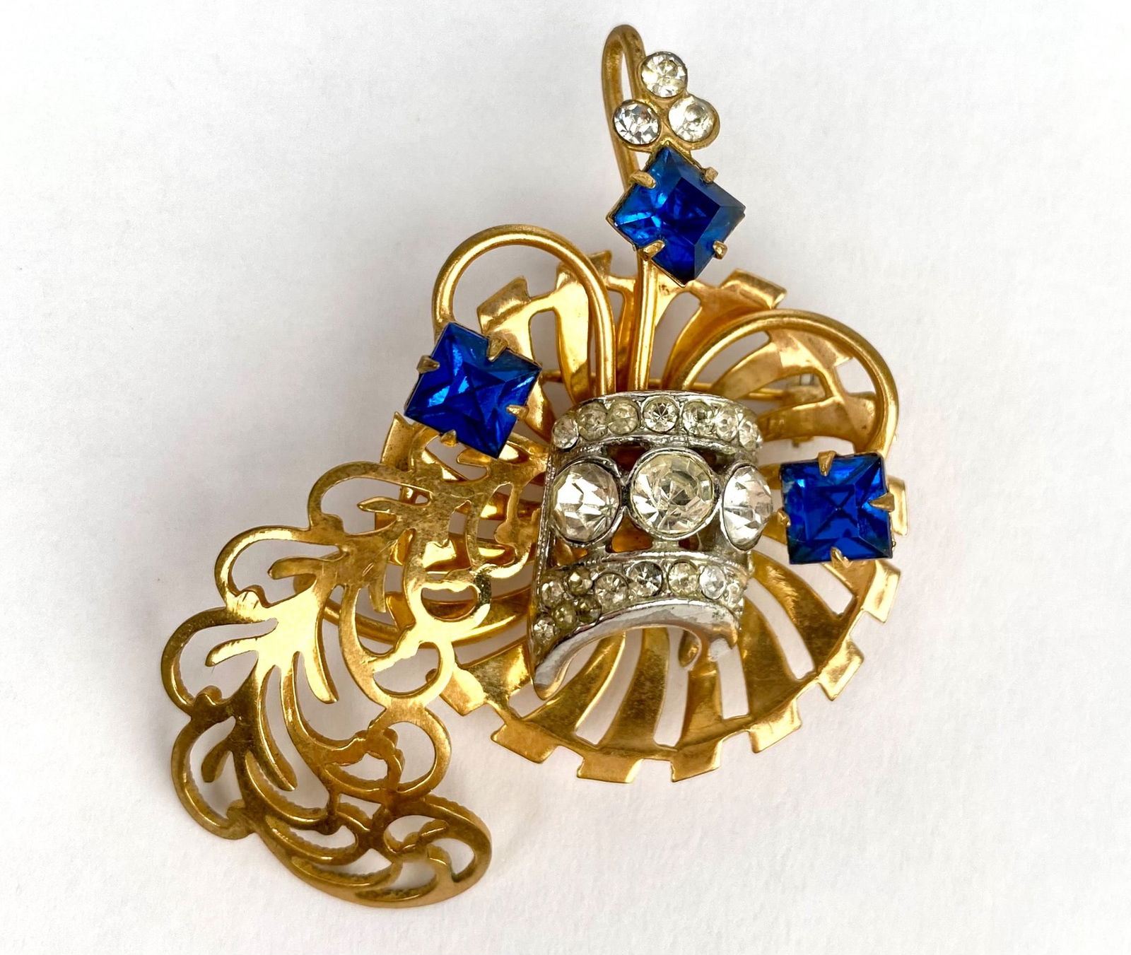 Retro Modern Costume Jewelry Brooch: Gold tone brooch featuring an ornate openwork design adorned with sapphire blue and clear rhinestones in varied cuts. Brooch is unmarked but is consistent with brooch makers from the 1940s-50s. Item i