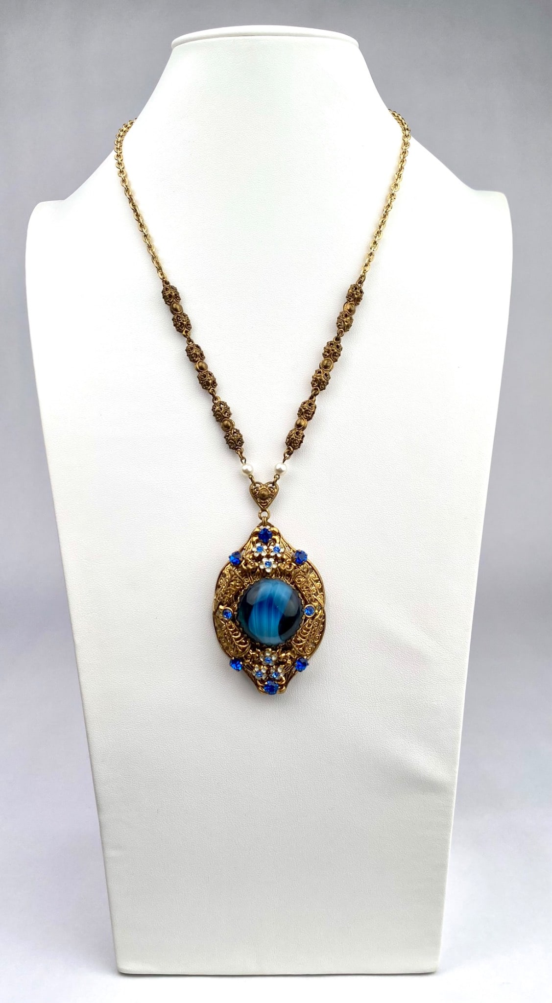 Antique West Germany Blue Glass and Rhinestone Necklace: Gold tone pendant necklace featuring a central large blue cabochon set within intricate filigree work. The pendant is adorned with blue and clear rhinestones. The chain features a combination of textu