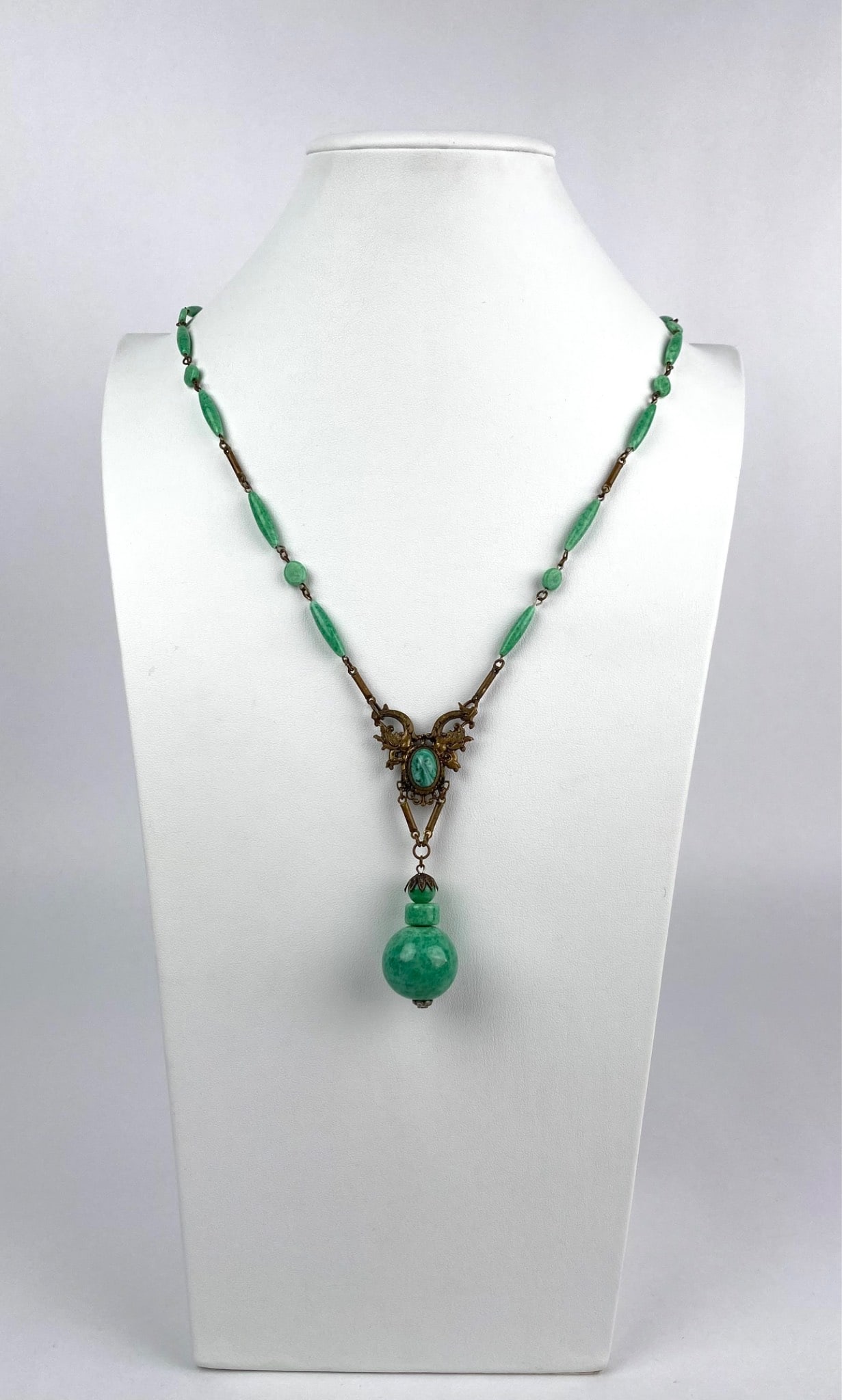 Vintage Art Nouveau Style Necklace with Green Glass Drop Pendant: Art Nouveau style necklace featuring a green faceted drop pendant suspended from an ornate brass tone centerpiece. The openwork setting showcases a bezel set marbled green cabochon, flanked by decorat