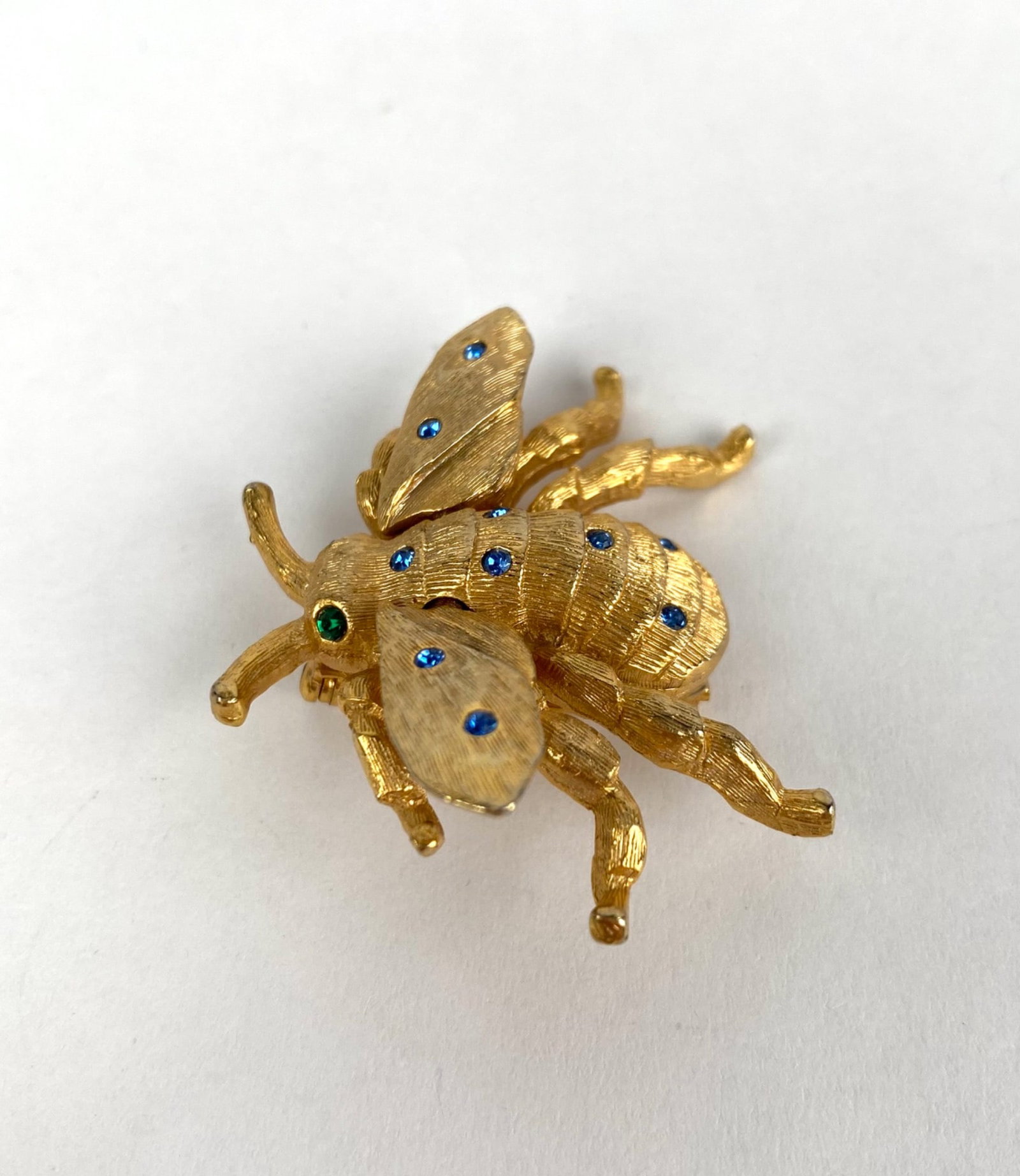 Denicola Gold Tone Bee Brooch Adorned With Rhinestones: Gold-tone brooch in the shape of a bee, designed by Denicola, featuring blue rhinestones on the wings and body, and a green rhinestone as one eye. The textured surface enhances its detailing. The back