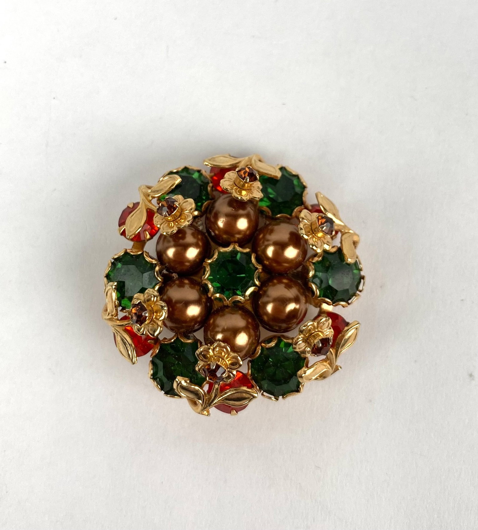 Gold Tone Brooch with Green and Red Rhinestones: A circular gold tone brooch featuring a detailed design incorporating green and red rhinestones, golden faux pearls, and floral decorative elements. The reverse showcases a secure pin mechanism for at