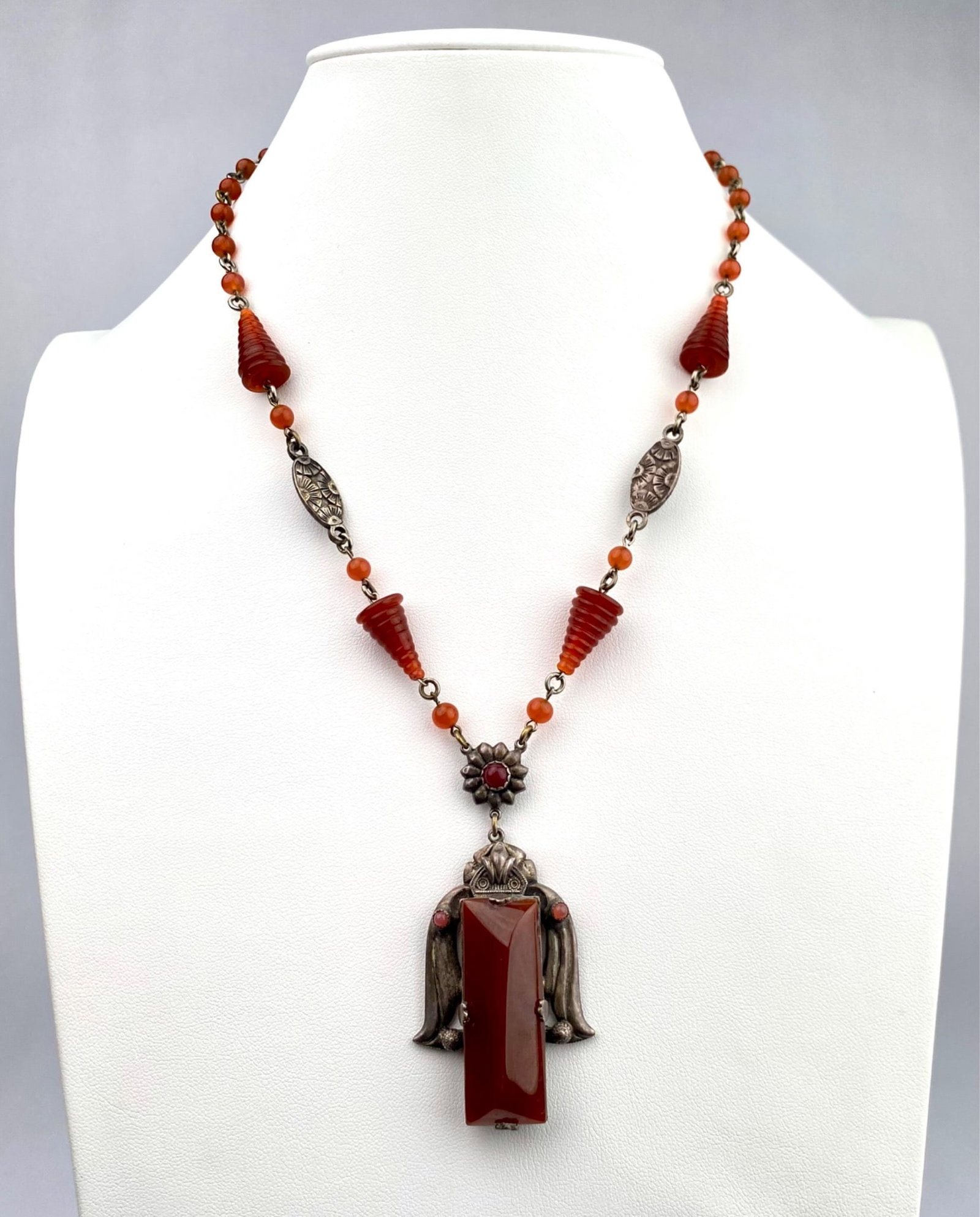 Silver Tone and Carnelian Pendant Necklace with Ornamental Beads: Silver and carnelian pendant necklace featuring a central drop design with detailed silverwork and a large faceted carnelian centerpiece. Accented by ornate cone shaped carnelian beads, round carnelia