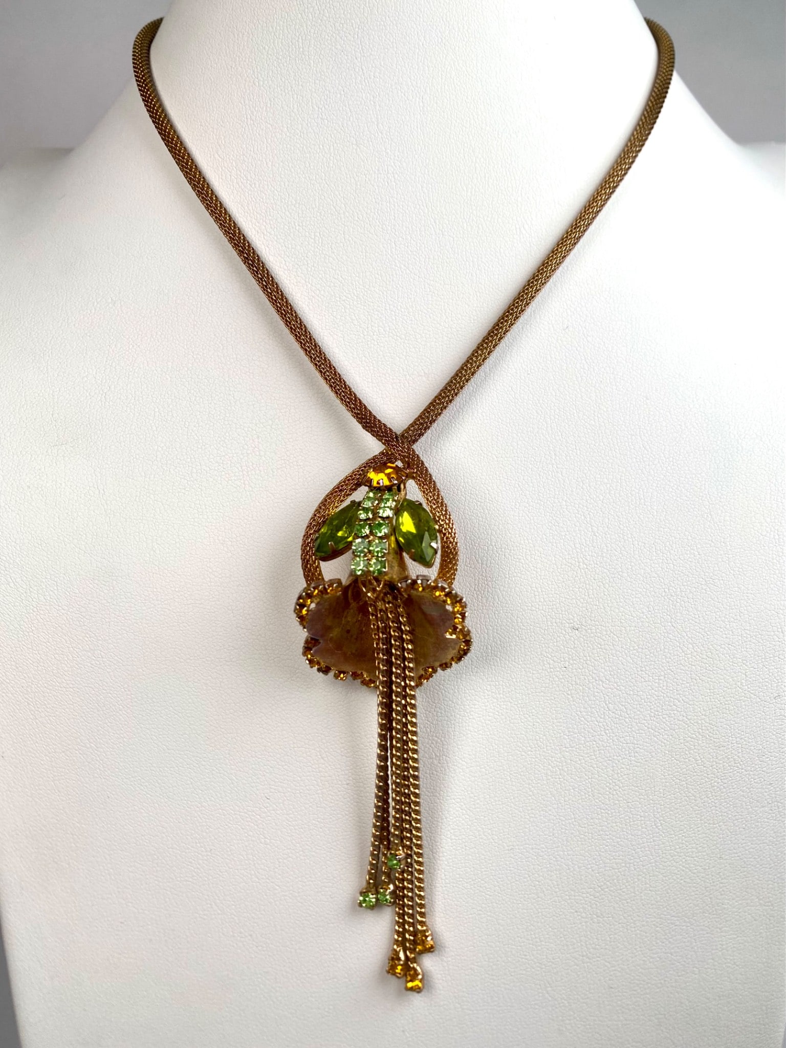 Necklace Featuring Green and Amber Rhinestone Floral Pendant: Gold tone necklace featuring a pendant in a floral design adorned with green and amber rhinestones. The pendant includes dangling chains accented with matching green and amber stones. It is suspended 