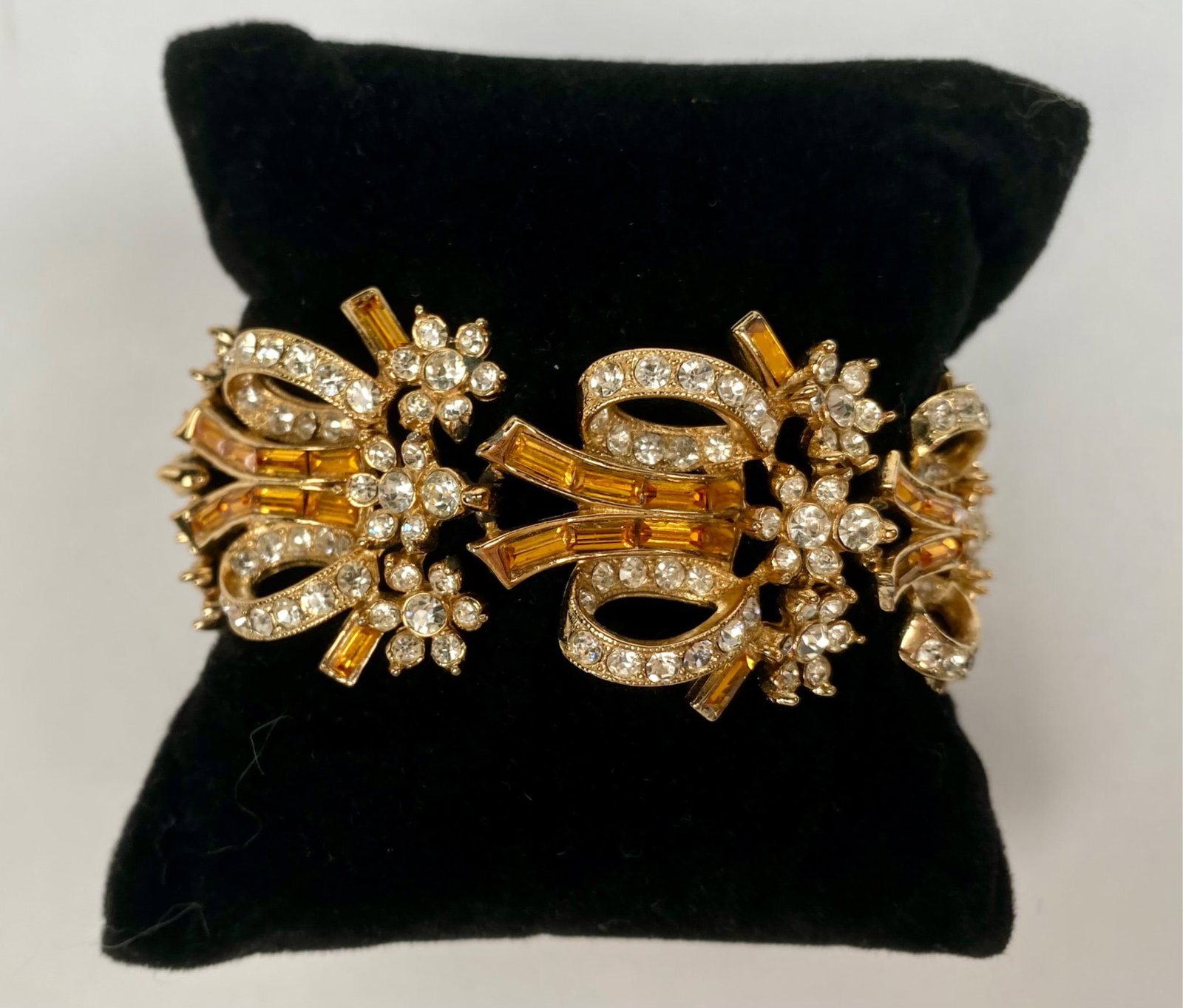 ORA Rhinestone and Glass Baguette Bracelet: Gold tone bracelet crafted by ORA, featuring clear round rhinestones and amber hued glass baguettes in an ornate bow inspired design. The bracelet is adorned with floral accents and includes a safety 