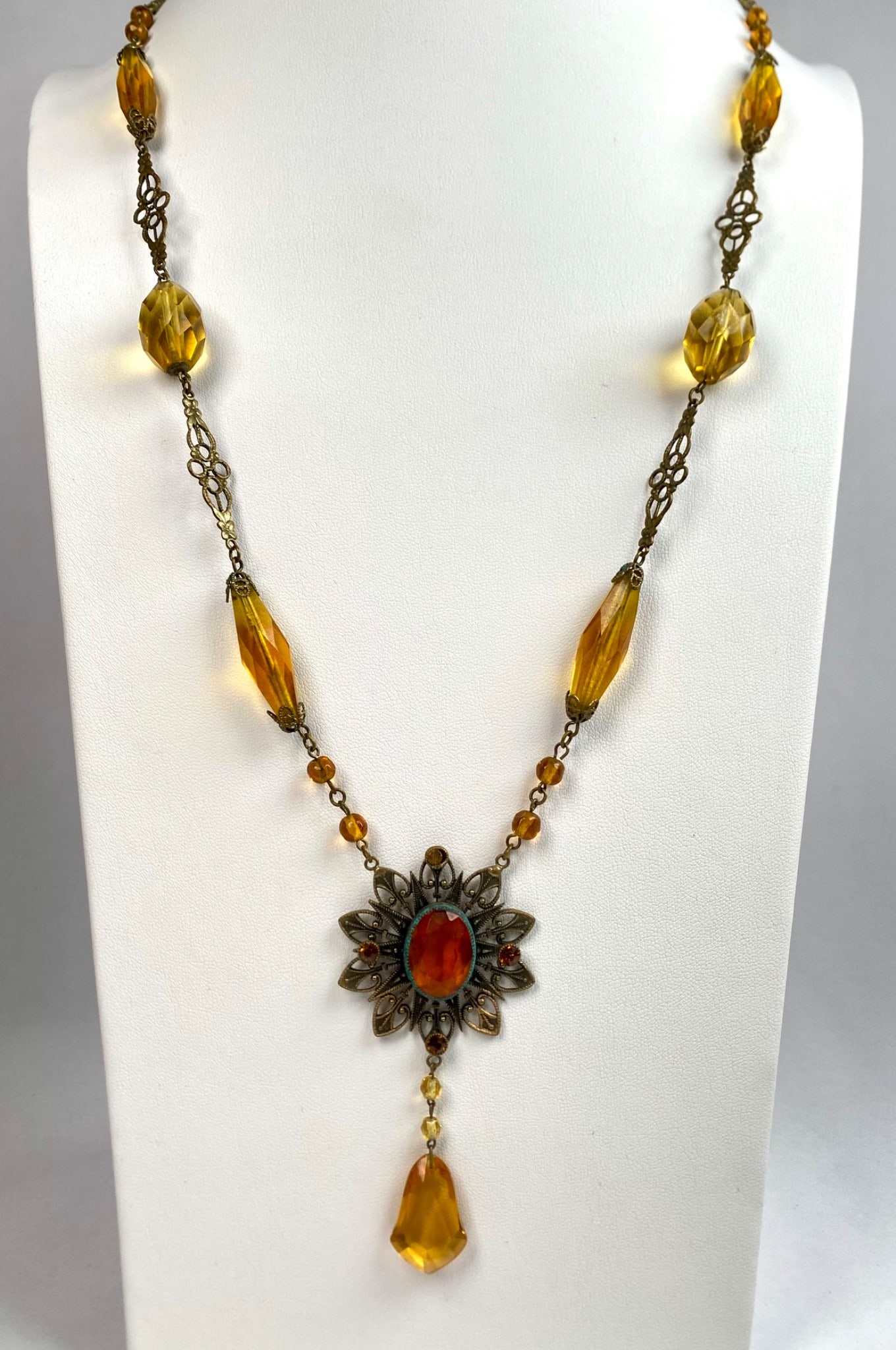Amber Glass and Filigree Pendant Necklace: Amber colored glass necklace featuring a filigree centerpiece with an ornate oval glass pendant surrounded by smaller matching rhinestone accents. The necklace includes elongated faceted glass beads, 