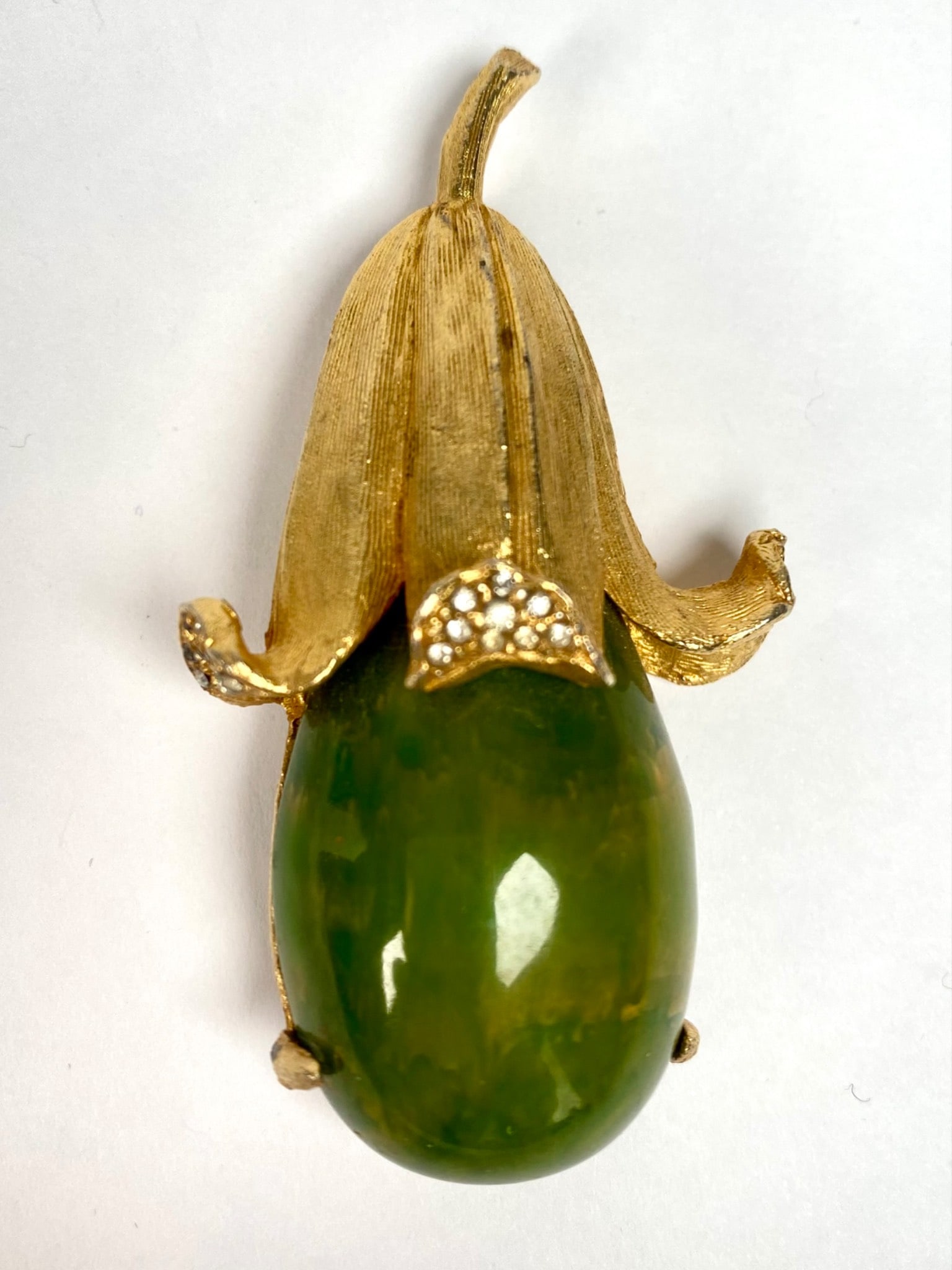 Green Stone Fruit Inspired Brooch Pendant with Rhinestone Accents: Gold tone brooch designed in a fruit motif featuring a green simulated jade stone centerpiece. The brooch is embellished with clear rhinestone accents for added detail. It includes a secure pin backin