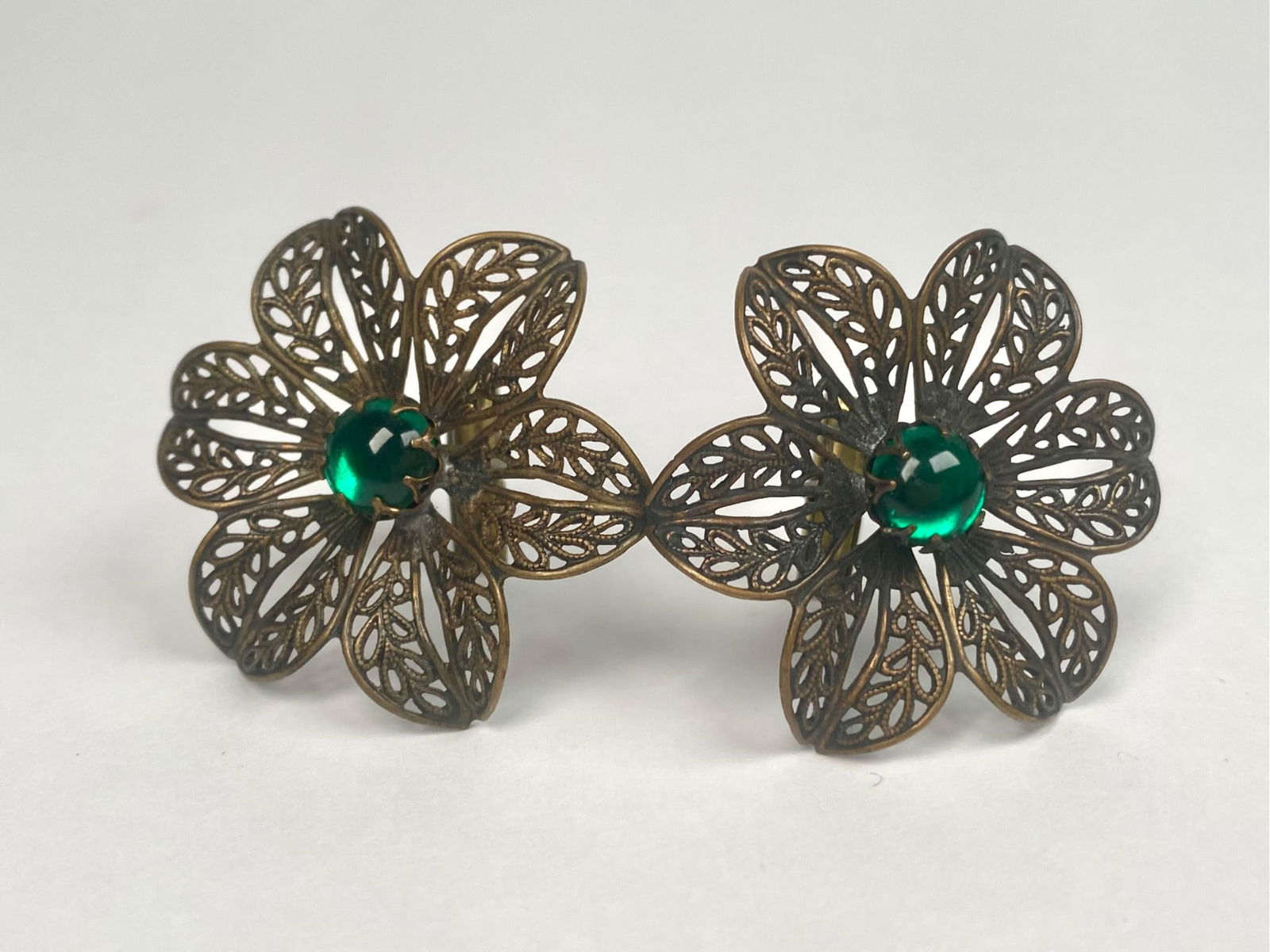 Pair of Brass Filigree Cuff Links with Green Glass Stones: A pair of vintage brass filigree cuff links featuring an intricate floral design. Each cuff link is adorned with a central green glass stone set within the center of the floral motif. The cuff links e