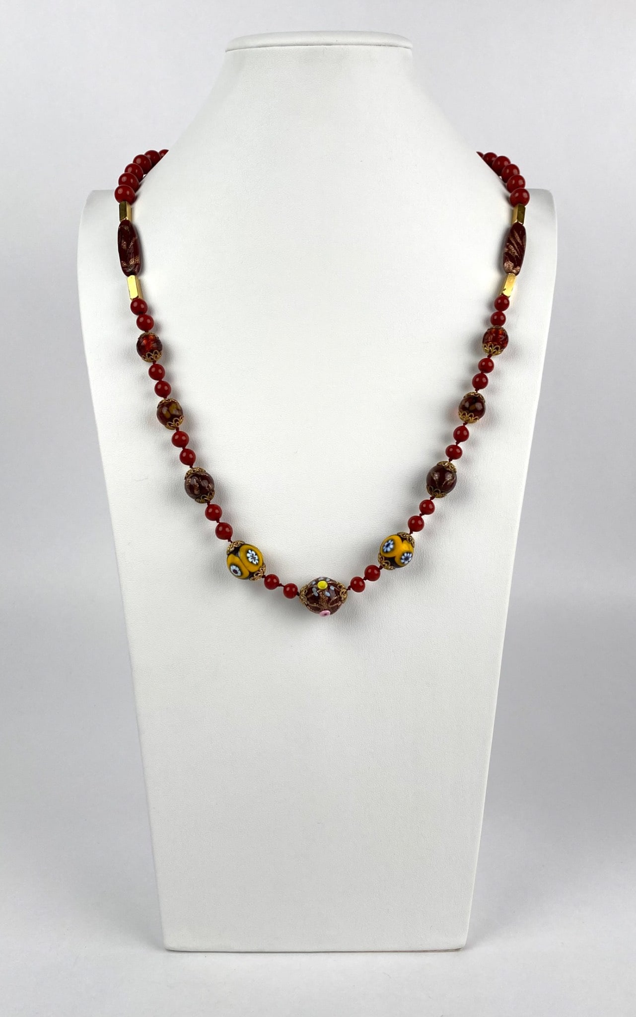 Glass Bead Necklace with Millefiori and Metal Accents: A handcrafted glass bead necklace featuring red beads interspersed with larger glass millefiori beads and intricate metallic accents. The centerpiece is composed of detailed millefiori work in yellow 