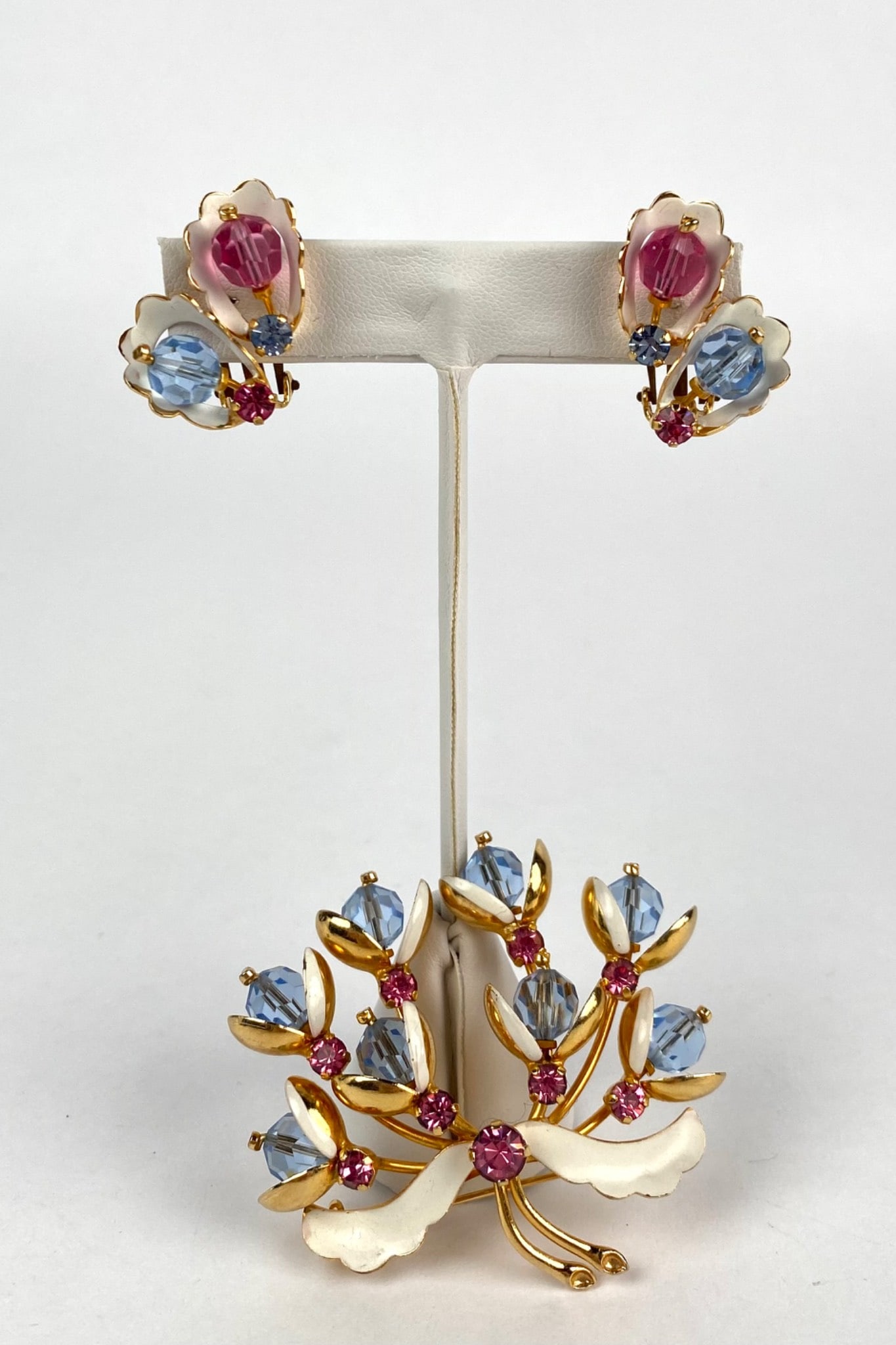 Floral Brooch and Clip On Earrings with Blue and Pink Crystal Accents: Floral brooch and matching clip-on earrings set crafted with gold-tone metal and adorned with blue faceted crystals, pink cabochons, and enamel detailing. The brooch features a floral spray motif with