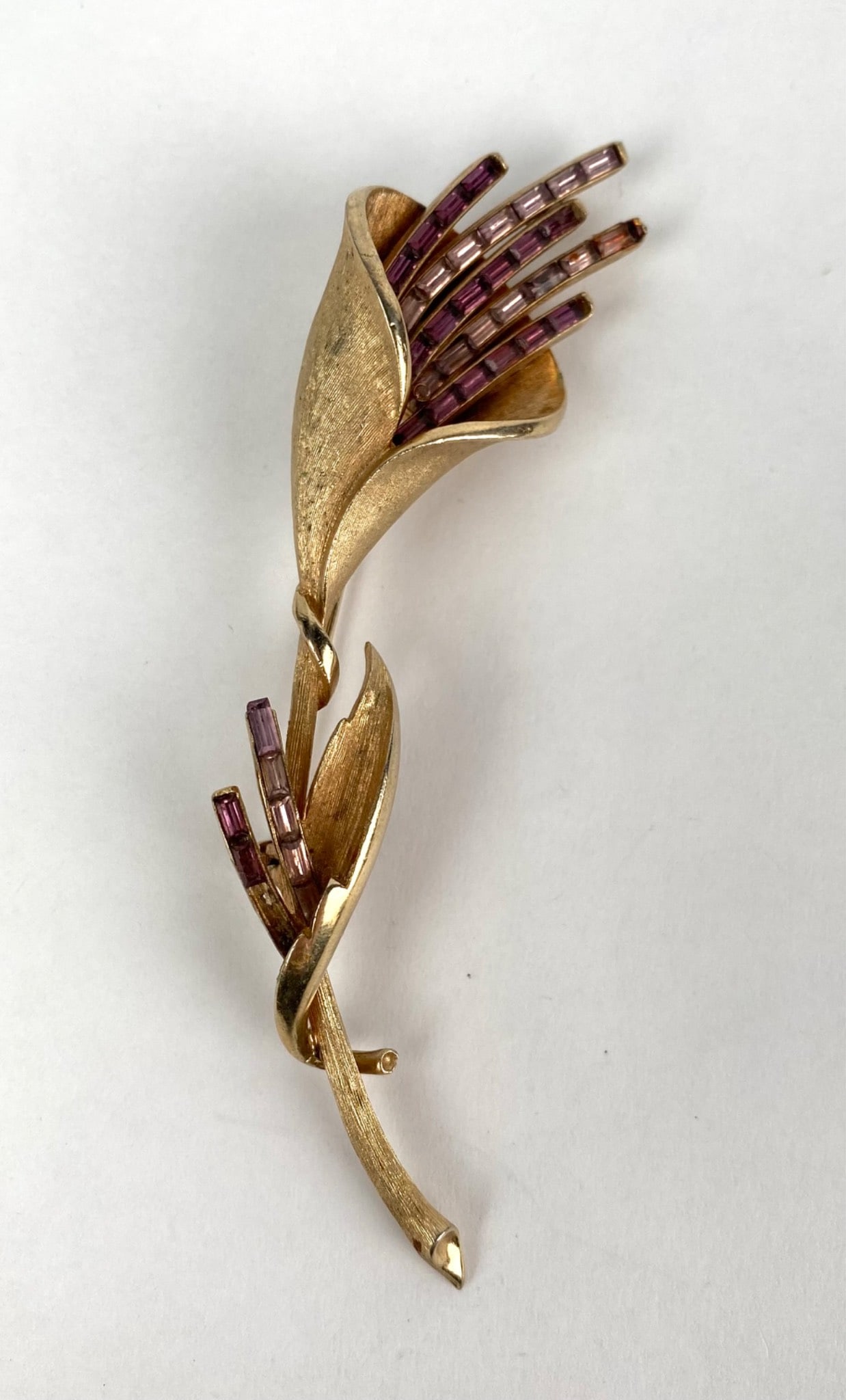 Coro Gold Tone Floral Brooch with Purple Crystal Accents: Gold tone brooch in a floral design featuring purple crystal baguette accents. Includes intricate texturing and sculptural details with a secure pin-back closure. Maker’s mark Coro visible on the re