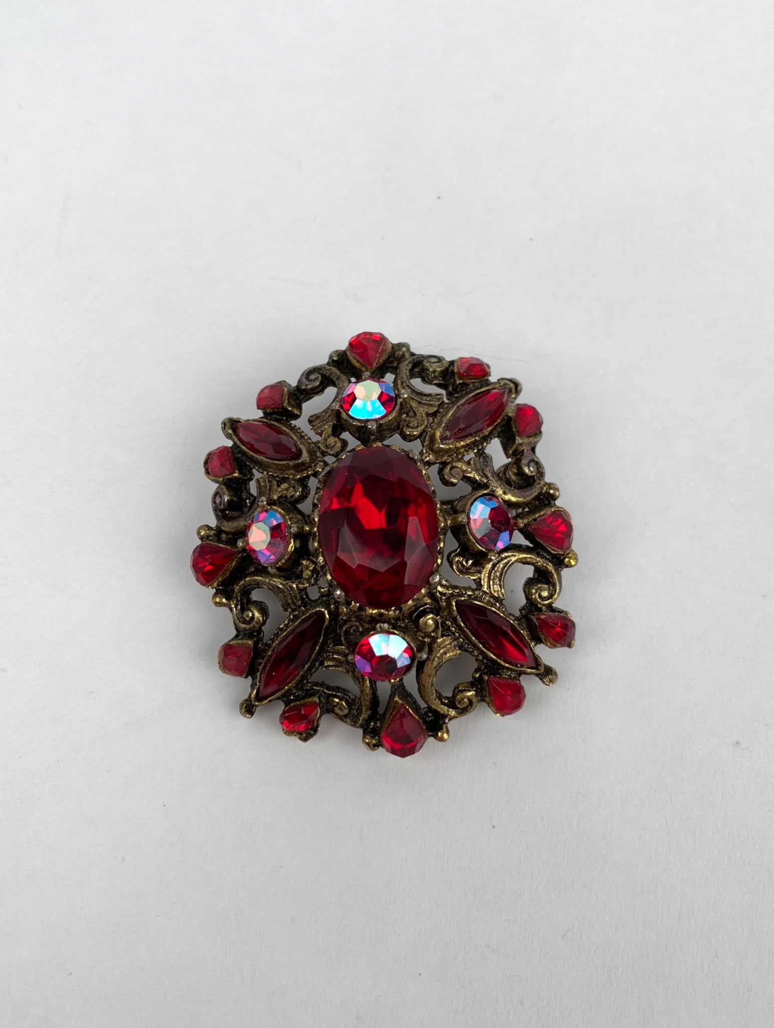 Vintage Style Gold Tone Brooch With Red and Iridescent Glass Stones: Gold tone metal brooch adorned with red glass stones and iridescent accent beads, featuring a central oval cut stone surrounded by ornate filigree detailing. The back includes a traditional pin clasp 