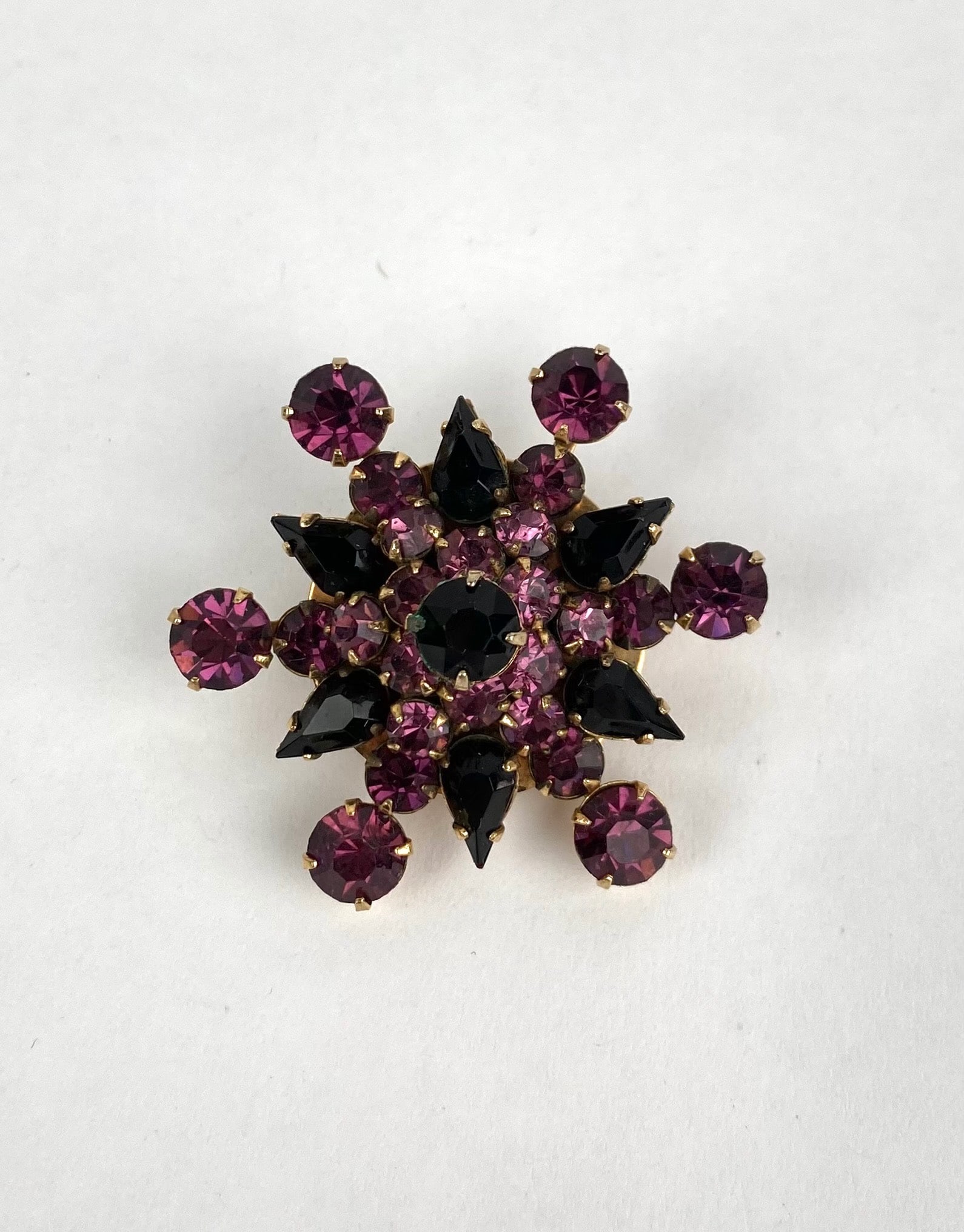 Gold Tone Floral Brooch with Purple and Black Rhinestones: Gold tone floral brooch featuring purple and black rhinestones in a dynamic, clustered design. The piece comprises both round and teardrop shaped stones, set in a layered arrangement. The back shows a