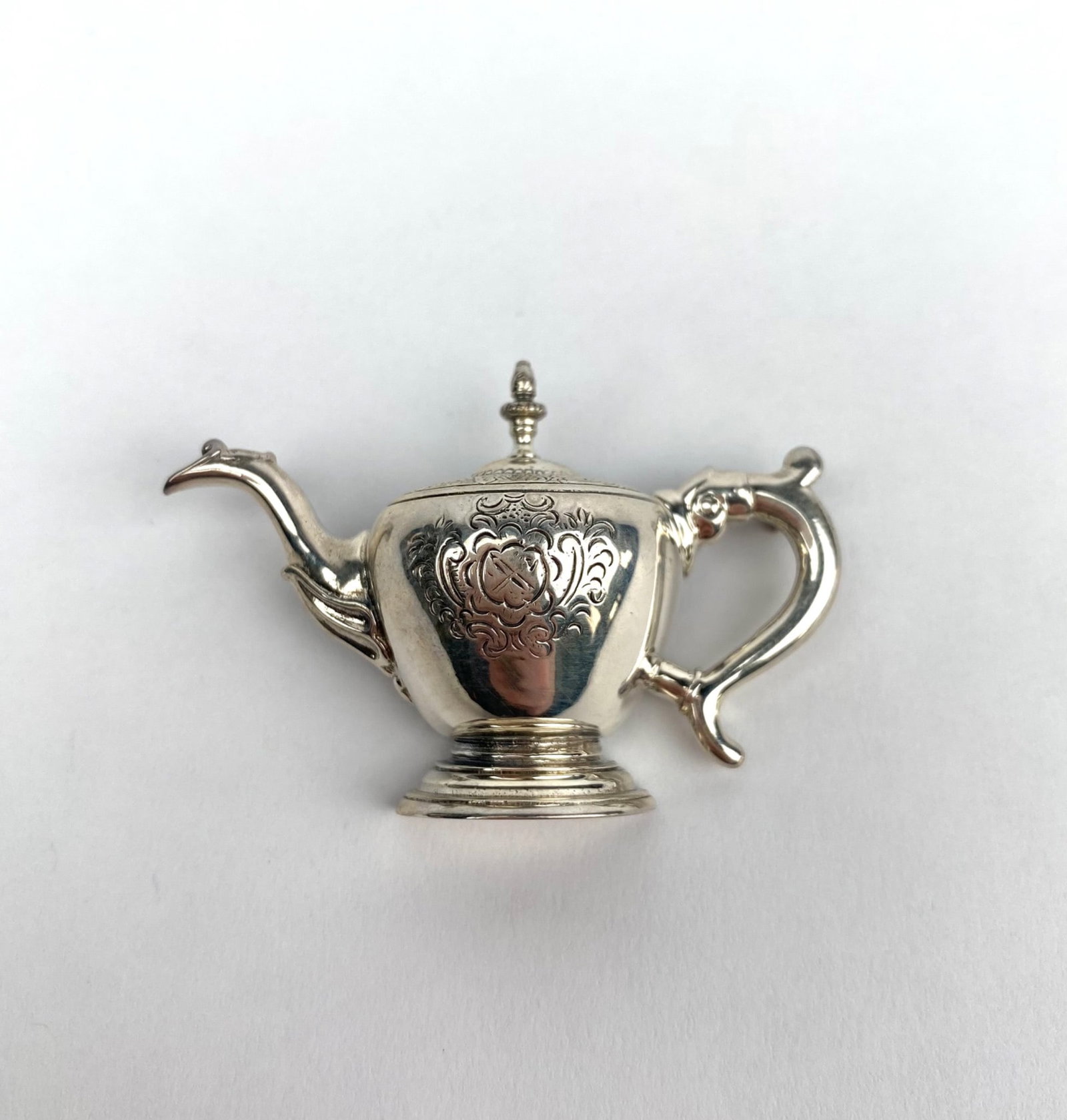 Teapot-Shaped Sterling Silver Brooch With Engraved Details: Silver teapot-shaped brooch with engraved detailing on the front. Back side exhibits a pin fastening mechanism. Marked with 925, denoting sterling silver purity. Features ornate floral patterns on the
