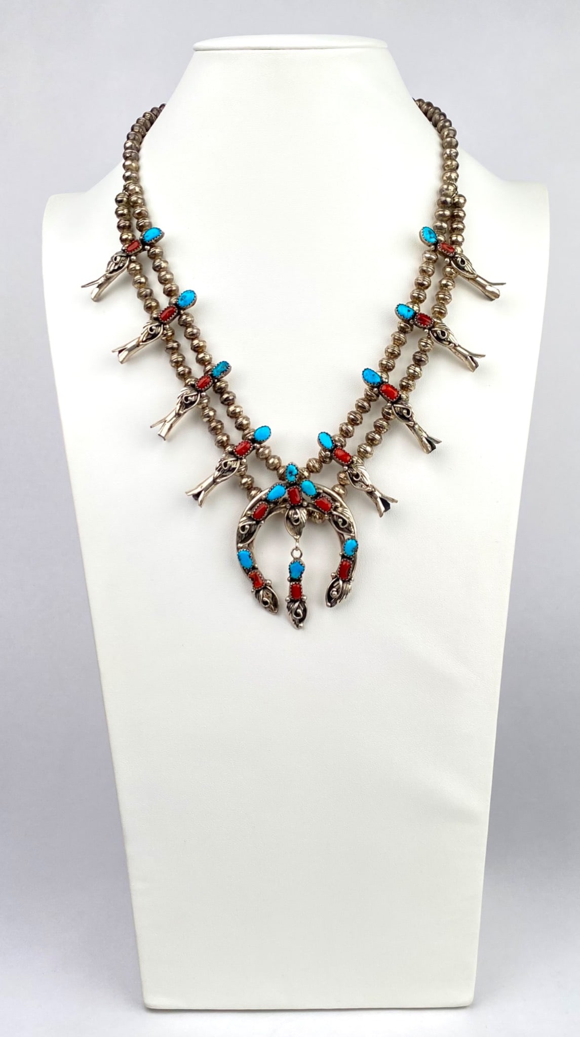Navajo Sterling Silver and Turquoise Squash Blossom Necklace: Sterling silver squash blossom necklace featuring turquoise and coral stone accents. The necklace consists of a double strand of sterling beads and showcases a naja pendant ornamented with turquoise a