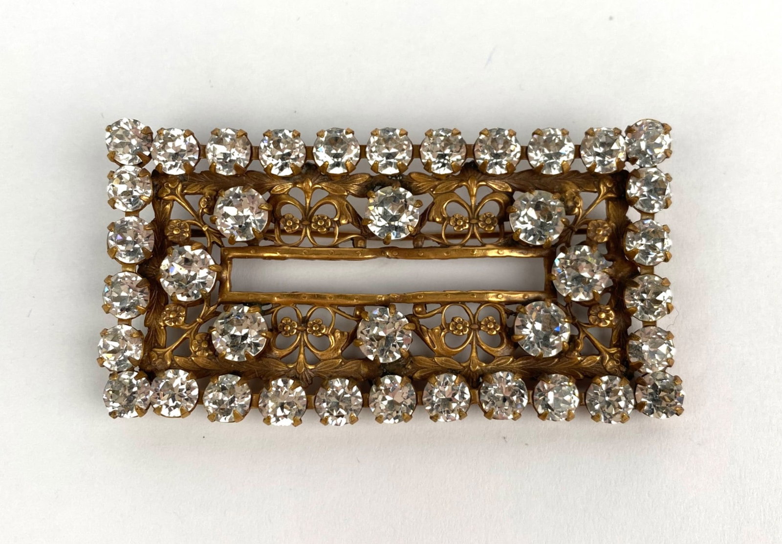 Vintage Rectangular Brooch With Rhinestones and Filigree Design: Gold-tone rectangular brooch adorned with clear rhinestones set in a double-row design. Features intricate filigree detailing and a functioning pin and clasp mechanism. A hallmark is visible on the ba