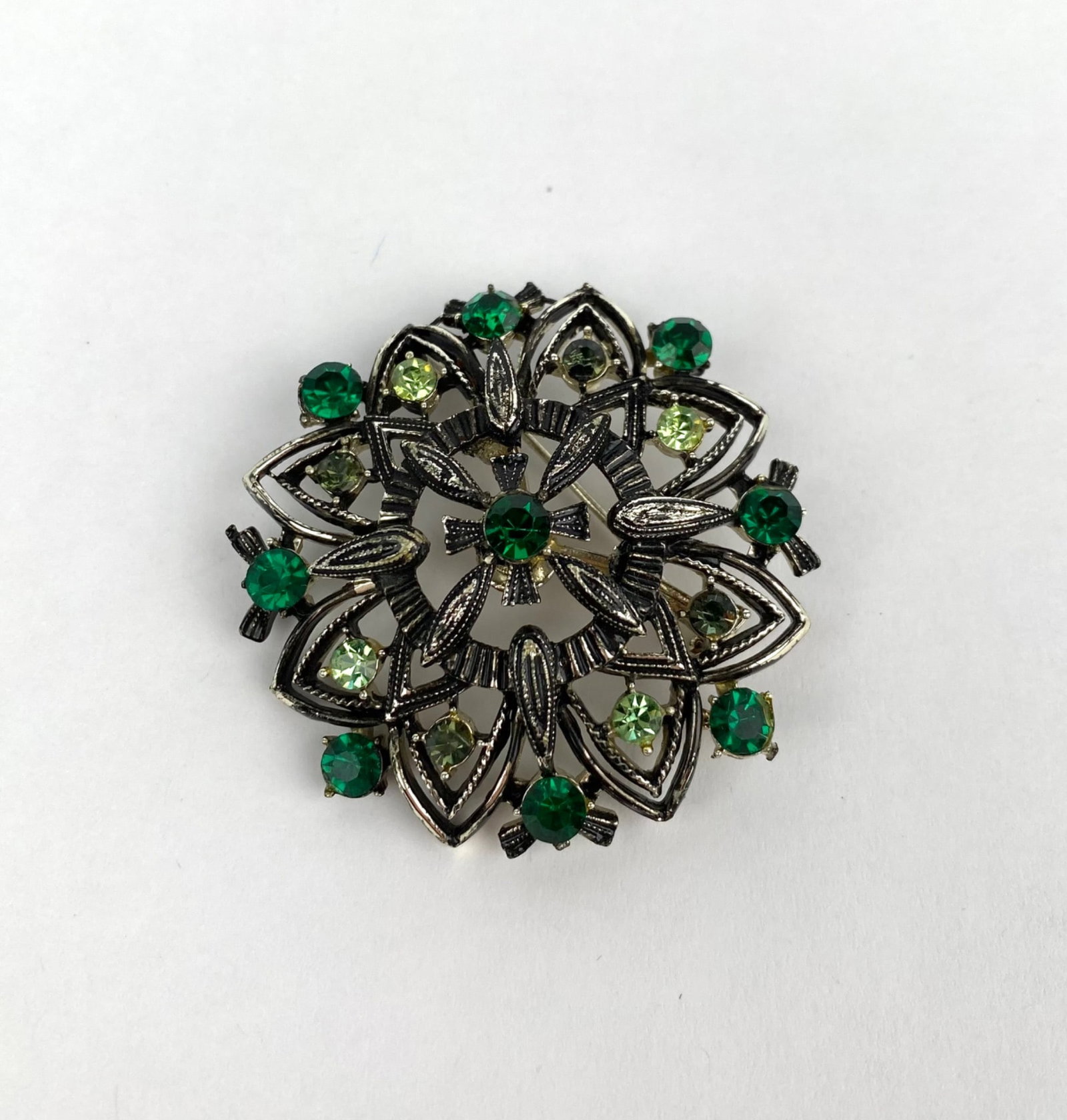Mid Century Floral Brooch: Silver tone floral brooch featuring an intricate overlay of pierced geometric patterns adorned with green and clear rhinestones. The back features a functional pin and clasp mechanism. In good conditi