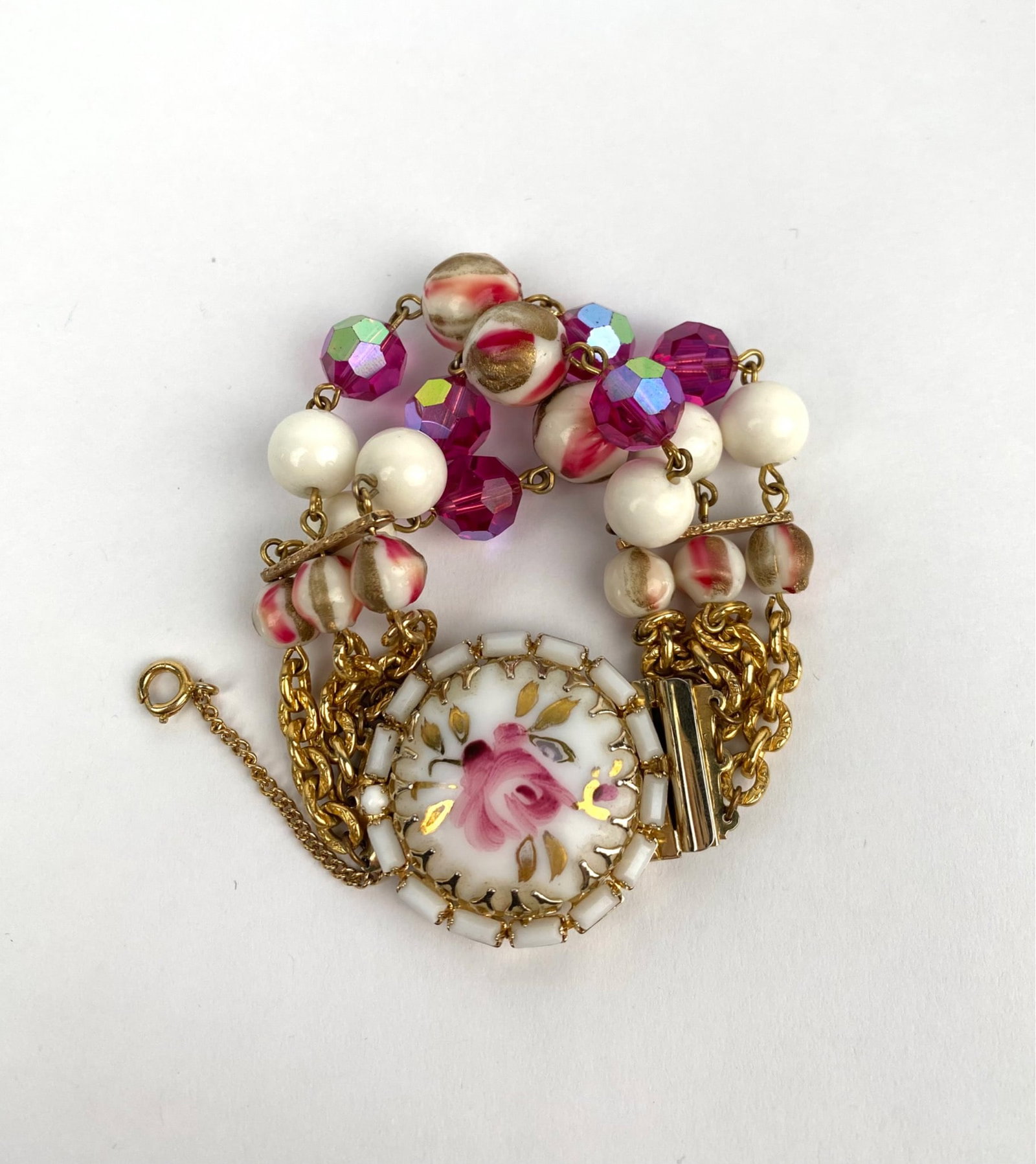 Hobe Mid Century Multi Strand Bracelet: Hobe gold tone bracelet featuring a painted pink rose on a circular clasp centerpiece and accented by multi strand chains. The bracelet is adorned with white beads, pink and rose patterned beads, and 