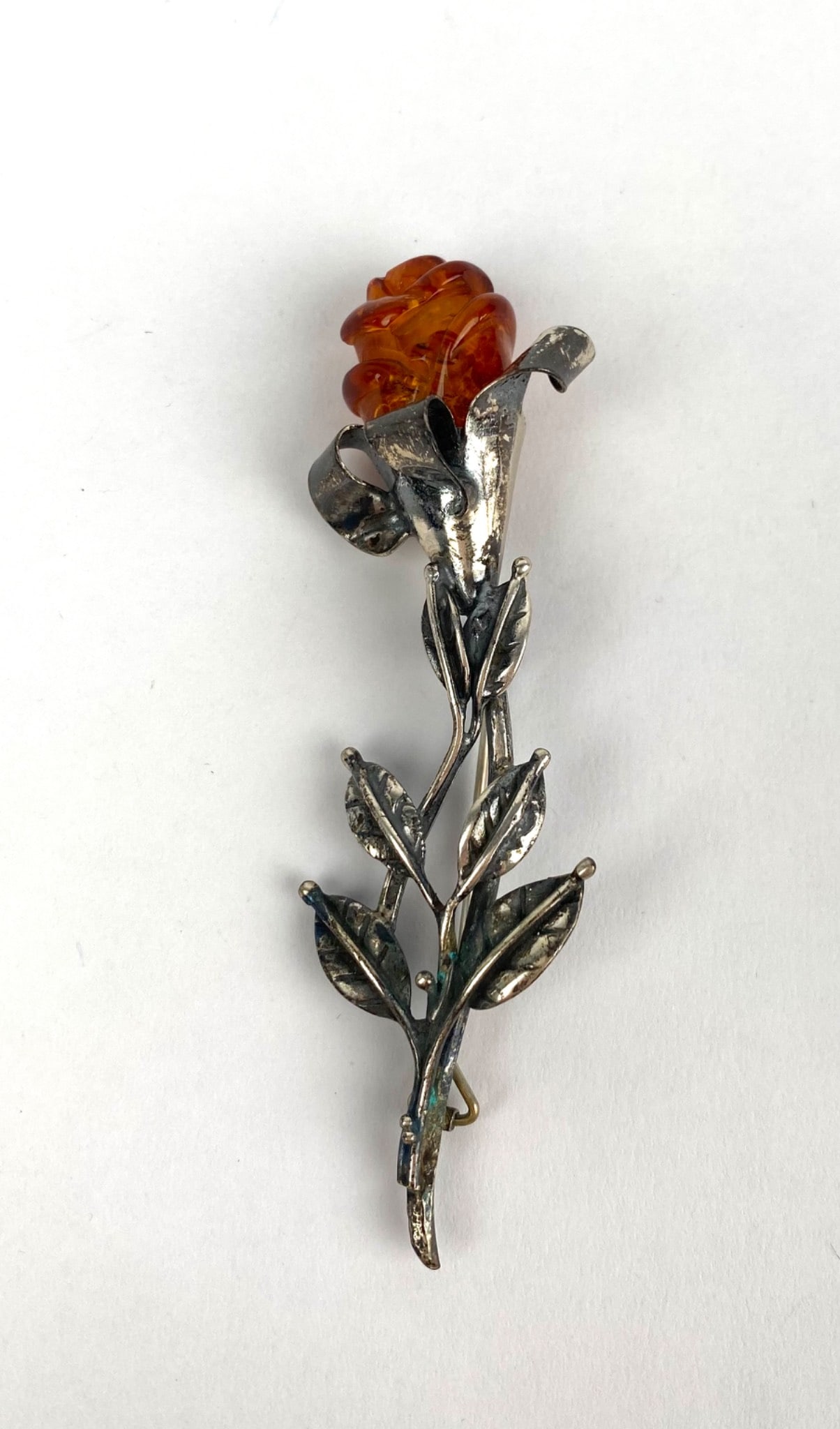 Vintage Amber Rose Form Brooch: Amber and silver brooch designed in the form of a rose. The piece features a carved amber rose as the focal point and a detailed silver stem and leaf pattern. The brooch includes a hinged pin closure.