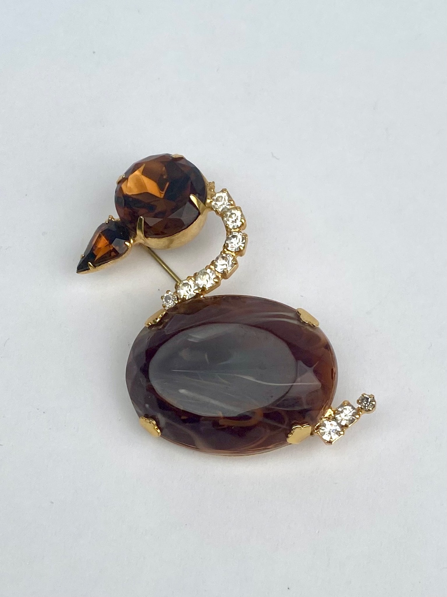 Swan Shaped Brooch with Rhinestone and Brown Glass Accents: Gold tone brooch featuring a large oval brown stone and round topaz colored rhinestones accented with a curved row of clear rhinestones and a small teardrop shaped element to create a beak. Designed i