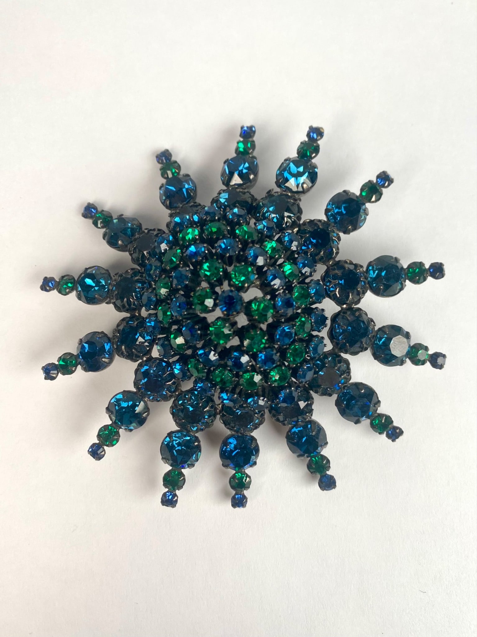 Mid Century Sunburst Pendant Brooch with Japanned Finish: Japanned finished costume brooch featuring a sunburst design with prong set blue and green faceted rhinestones. The vivid colors of the blue and green stones creates a dazzling contrast against the da