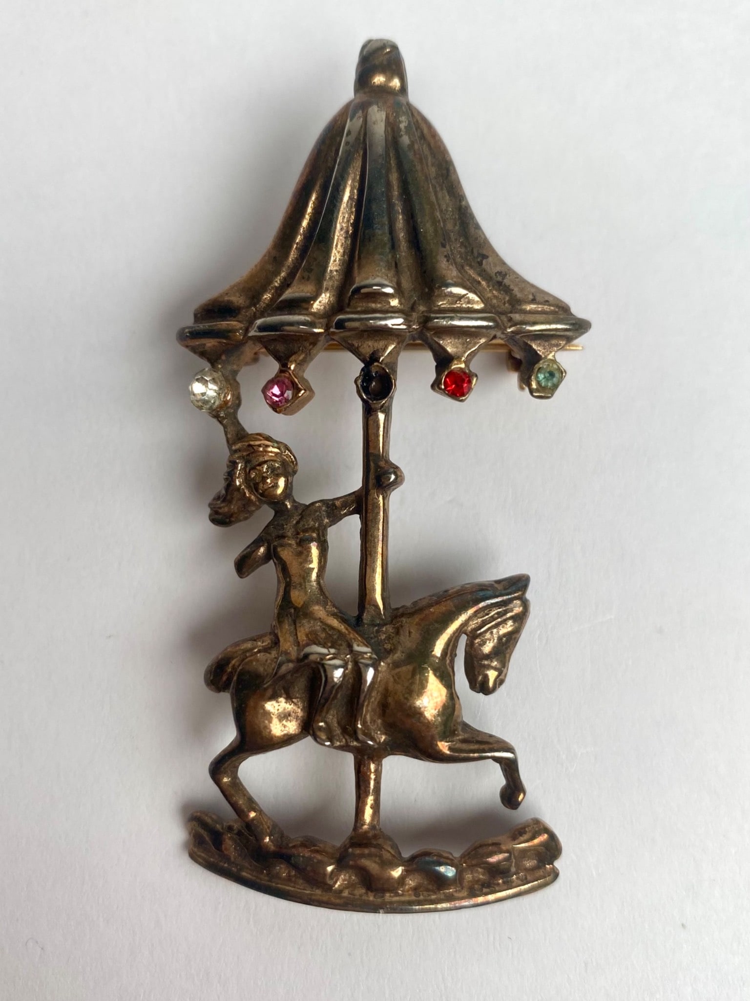 Vintage Sterling Silver Carousel Brooch: Sterling Silver brooch depicting a carousel horse and rider under a draped canopy, adorned with colorful gem accents, including red, green, white, and pink stones to mimic carnival lights. Intricate d