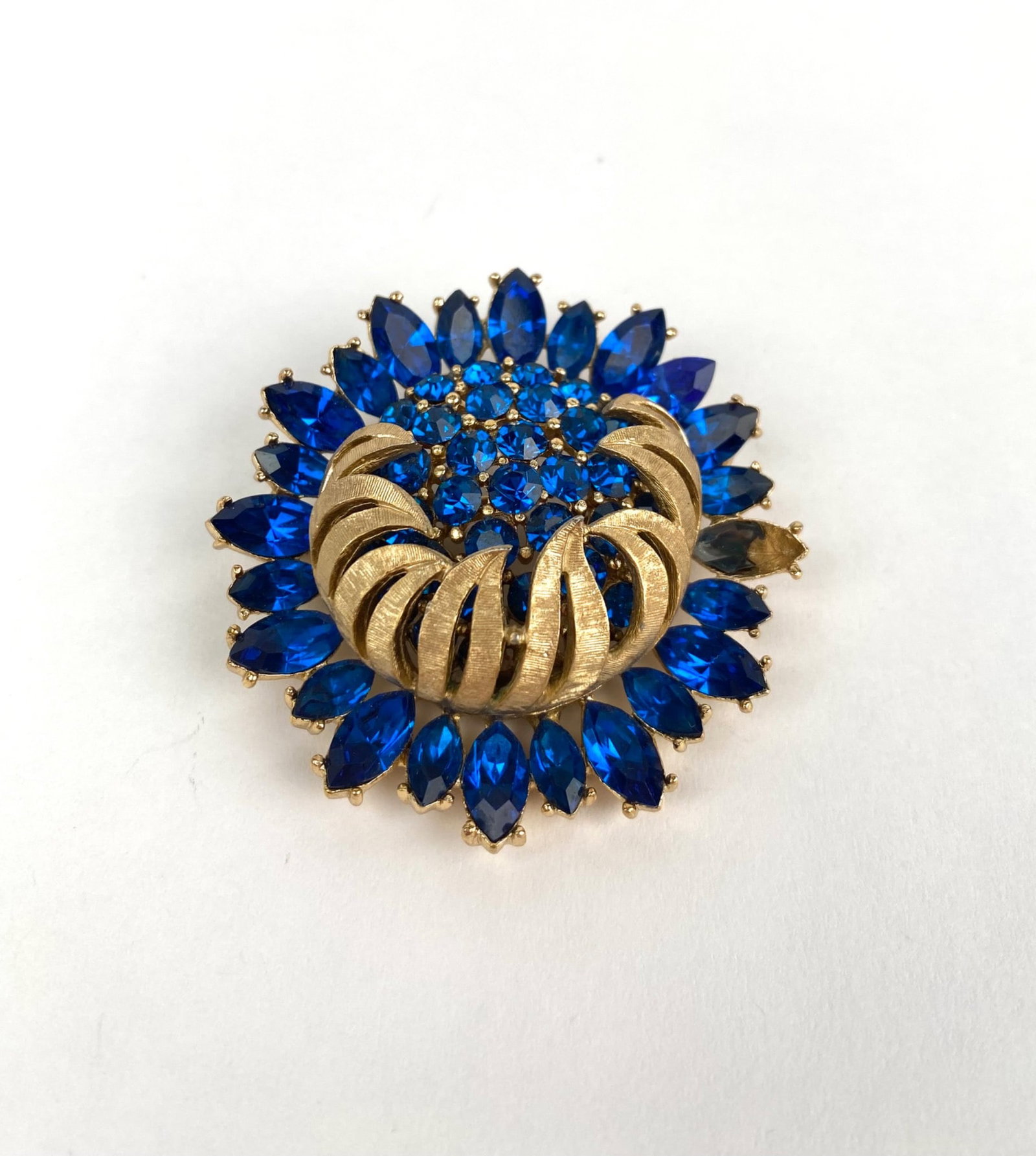 Crown Trifari 1940 to 1955 Gold Tone and Blue Rhinestone Brooch: Trifari brooch featuring gold tone metalwork and vibrant blue rhinestones in a layered floral design. The round brooch is set with marquise cut and round rhinestones, with a textured gold tone ribbon