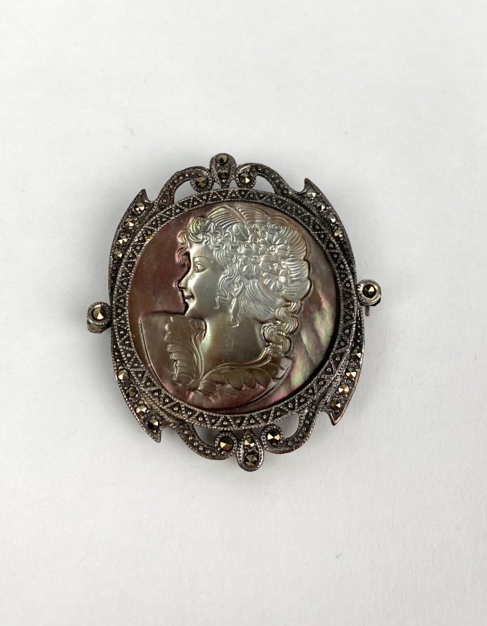 Mother of Pearl Mid Century Cameo Brooch: Mother of Pearl cameo brooch or pendant featuring a profile of a woman carved in relief. Framed with a marcasite accented ornate border. The piece includes a pin back and a loop for conversion into a