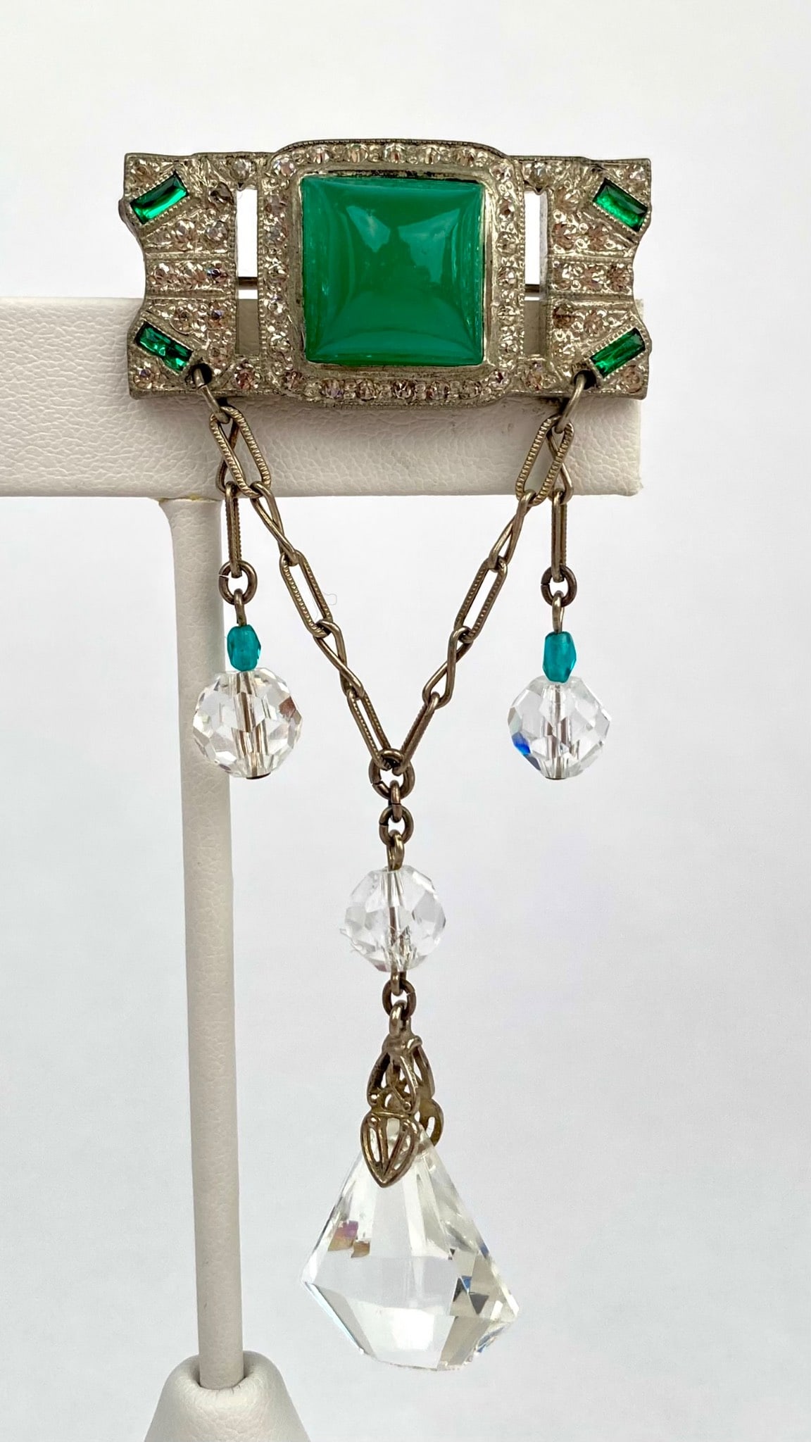 Art Deco Brooch with Green Glass and Crystal Drops: Art Deco style brooch featuring a central square green glass set in a silver tone metal frame, accented with green baguette and clear round stones. Two delicate chain drops are adorned with clear and