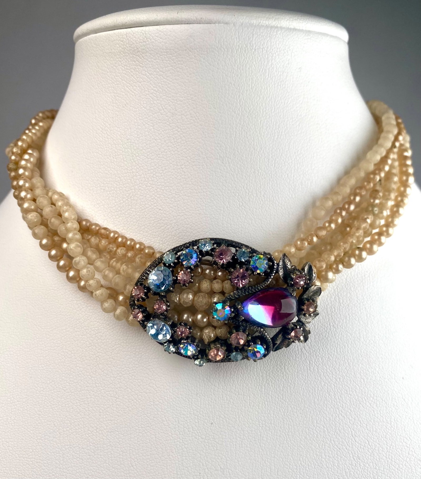 Vintage Multi Strand Faux Pearl and Rhinestone Choker: Vintage choker style necklace featuring six graduated strands of cream colored faux pearls with an ornate oval center piece with multi color rhinestones and a central magenta glass teardrop cabochon. 
