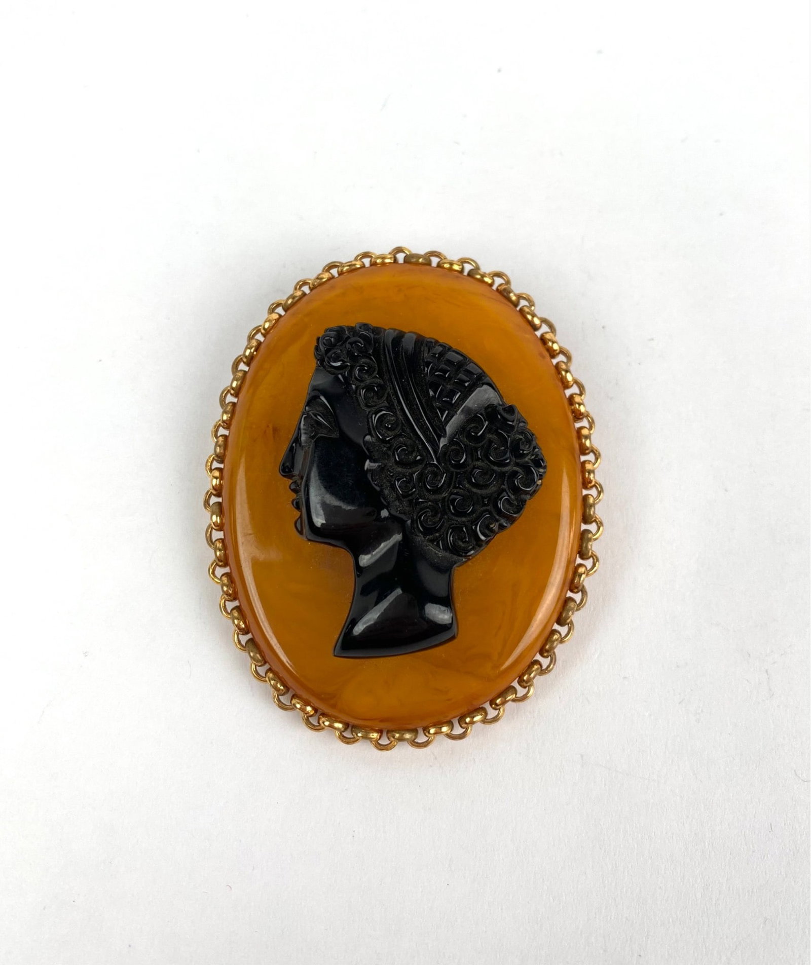 Vintage Amber and Onyx Cameo Brooch: Amber colored cameo brooch featuring a black carved profile of a female figure in high relief, framed in a gold tone chain like setting. The back is fitted with a functional pin for secure attachment.