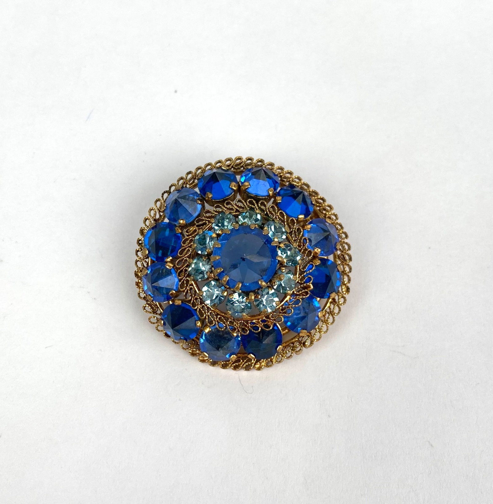 Vintage Austria Blue Rhinestone Gold Tone Filigree Brooch: A gold-tone filigree brooch featuring blue rhinestones in various sizes, arranged in a layered circular design. Marked Made in Austria on the back. Features a hinged pin and safety clasp for secure we