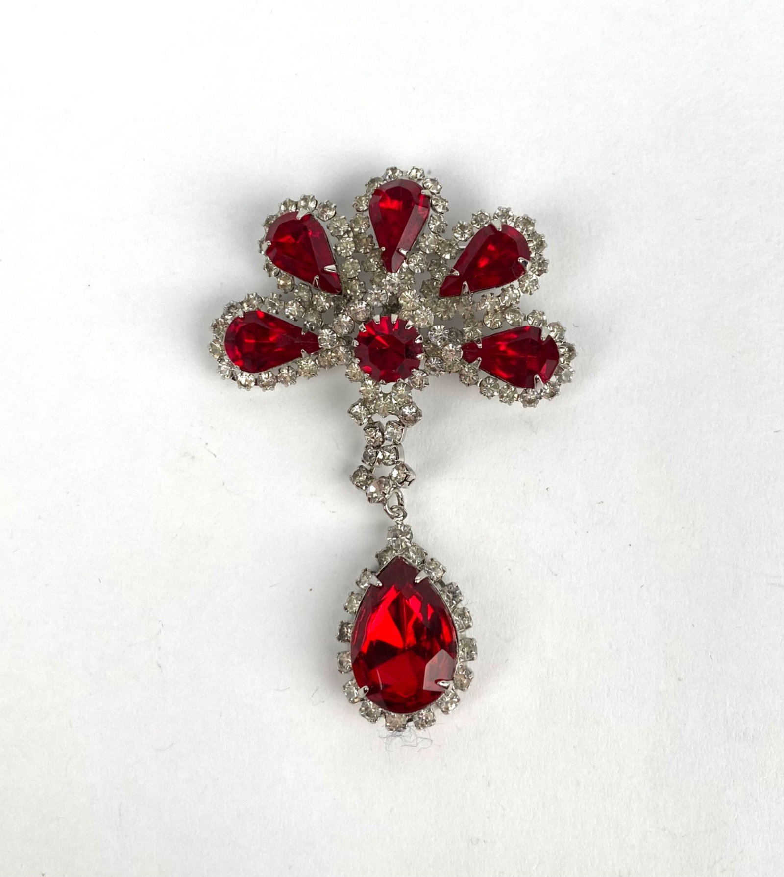 Mid Century Silver Tone Brooch With Red and Clear Rhinestones: Silver tone brooch featuring multiple red rhinestones in pear and round cuts, surrounded by clear rhinestones. The piece includes a dangling tear shaped red rhinestone detail, further accented by clea