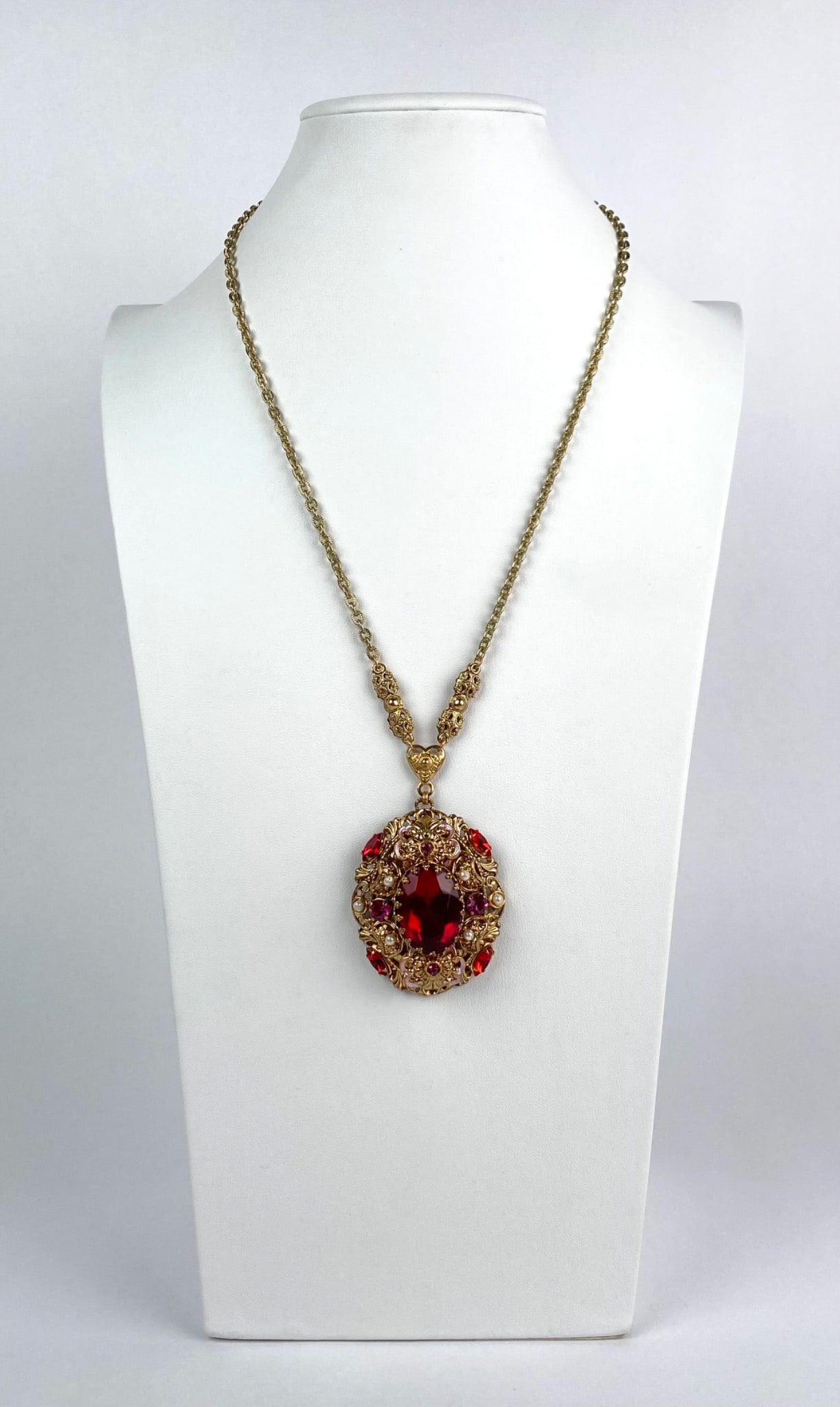 Western Germany Gold Tone Necklace Ornate Oval Pendent: Gold-tone chain necklace featuring an ornate oval pendant adorned with red and pink glass stones and faux pearls. The chain and pendant exhibit intricate detailing, with a secure clasp marked Western
