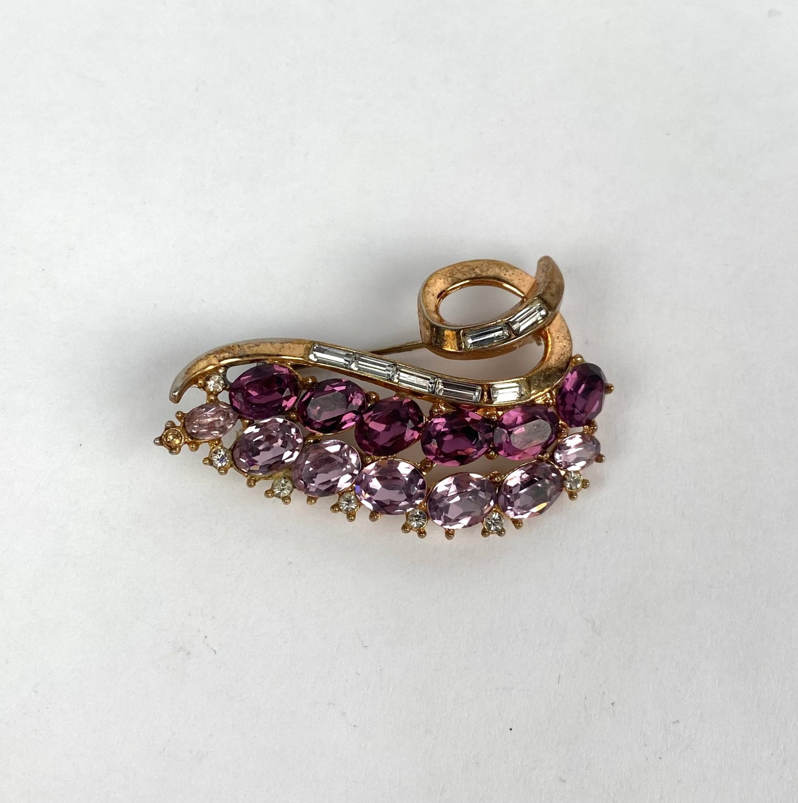 Trifari Gold Tone Brooch with Purple and Clear Rhinestones: Trifari gold tone brooch featuring purple and pink oval cut rhinestones arranged in rows with clear baguette cut rhinestones accenting the swirl design. The brooch is designed with ornate gold tone me