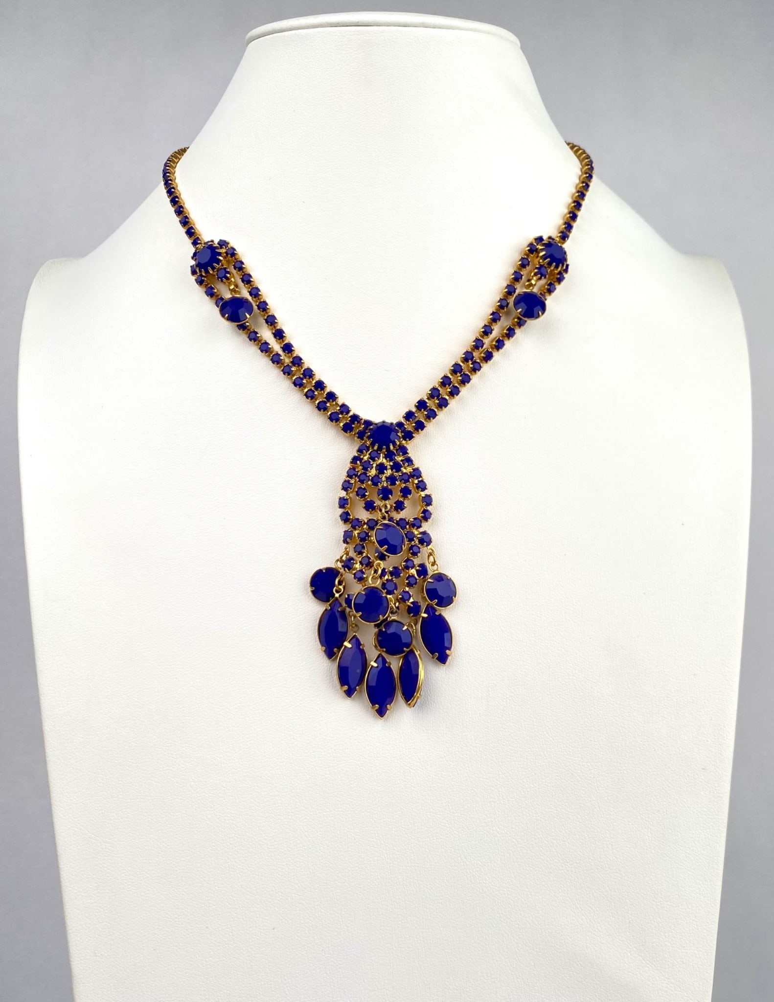 Gold Tone Necklace with Blue Glass Stones and Drop Pendant: Mid Century gold tone necklace featuring deep blue prong set glass stones arranged in a linked design, culminating in an elegant drop pendant with varying stone shapes. Secure lobster clasp closure at