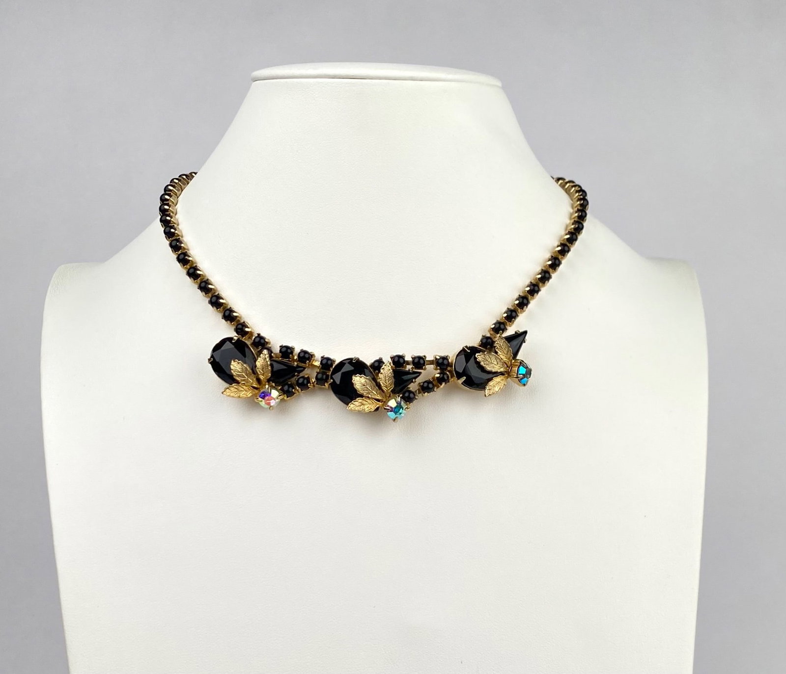 Mid Century Rhinestone Gold Toned Choker Necklace: Gold toned necklace featuring prong set black rhinestones along the length and drop elements adorned with faceted black stones, shimmering Aurora Borealis accents, and gold toned leaf motifs. Secures 
