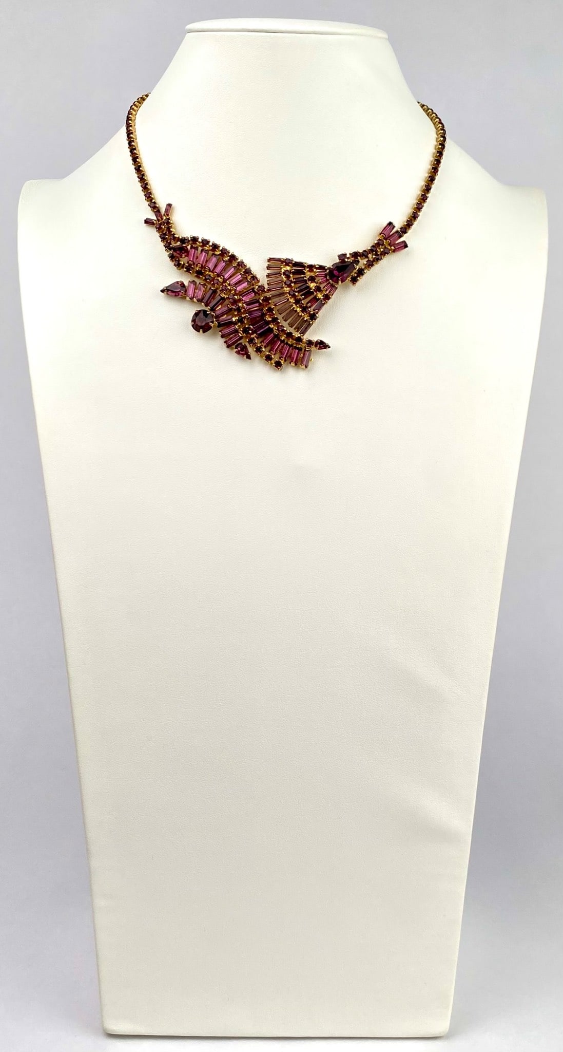 Juliana by DeLizza & Elster Amethyst Rhinestone Necklace: Gold toned necklace featuring a bold abstract design adorned with purple rhinestones in various shapes, including baguette cuts and round stones. The intricate design spans the front of the necklace, 