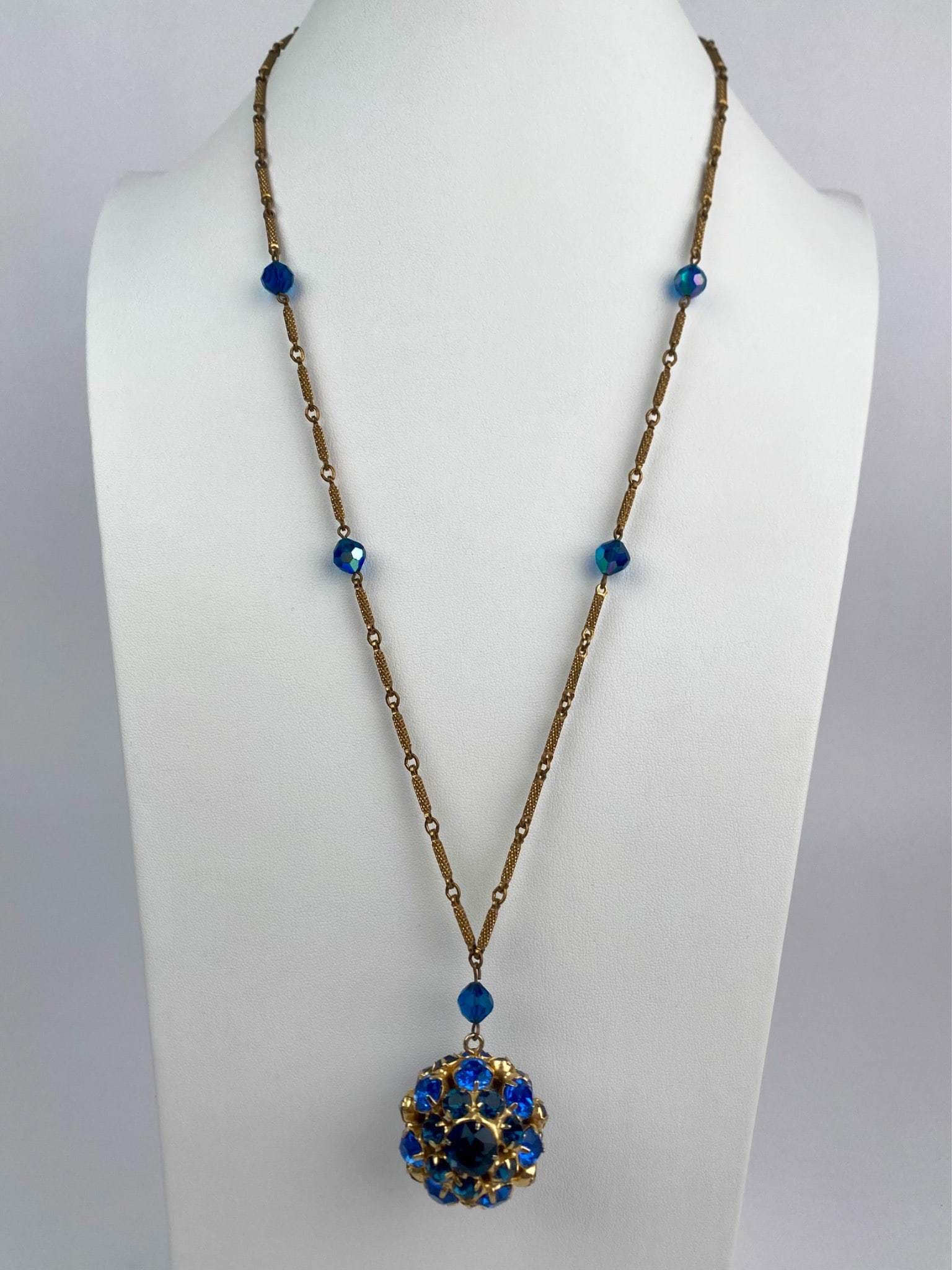 Blue Rhinestone Cluster Pendant Necklace on Textured Link Chain: Vintage necklace featuring a large domed pendant encrusted with vibrant blue rhinestones in a raised floral style cluster setting. The pendant is suspended from a finely textured gold tone bar link ch