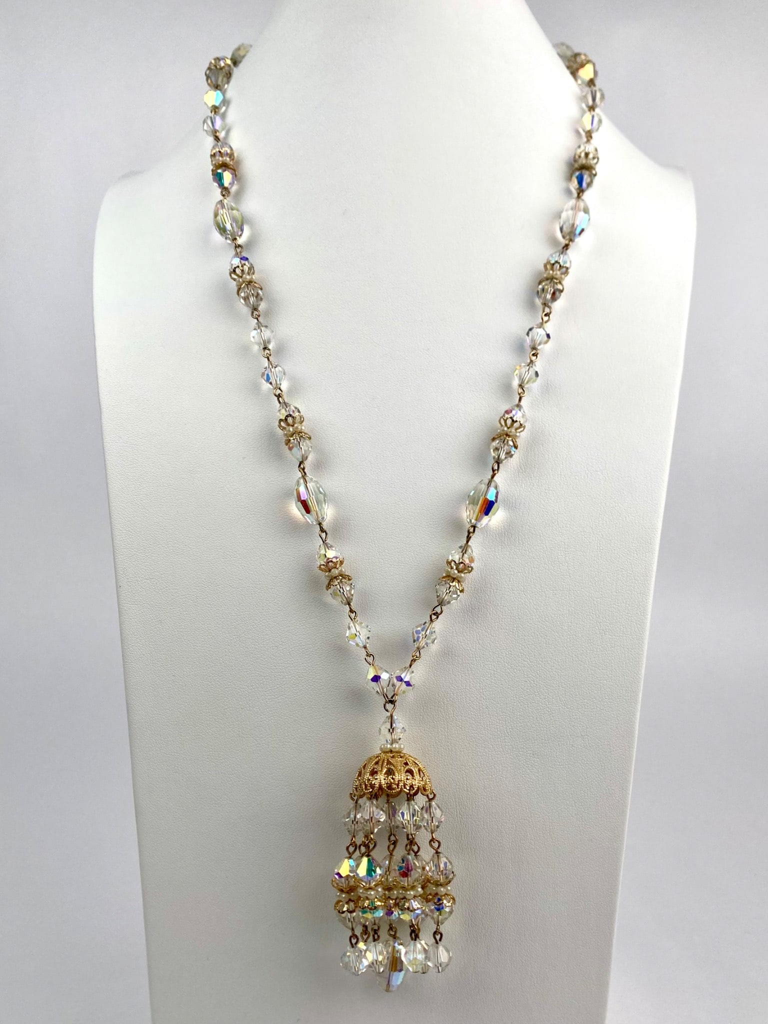 Vintage Aurora Borealis Crystal Tassel Pendant Necklace: Vintage necklace composed of faceted aurora borealis crystals, each bead shimmering with iridescent hues of gold, lavender, and blue. The strand alternating round and oval crystals with ornate faux pe