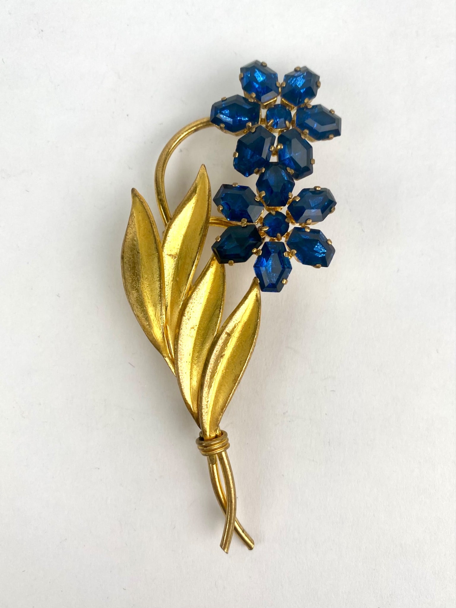 Vintage Czechoslovakia Blue Rhinestone Flower Brooch: Vintage floral brooch featuring two stylized flower clusters composed of rich sapphire blue faceted glass stones prong set in a bright gold tone setting. The flowers are accented by three curved gold 