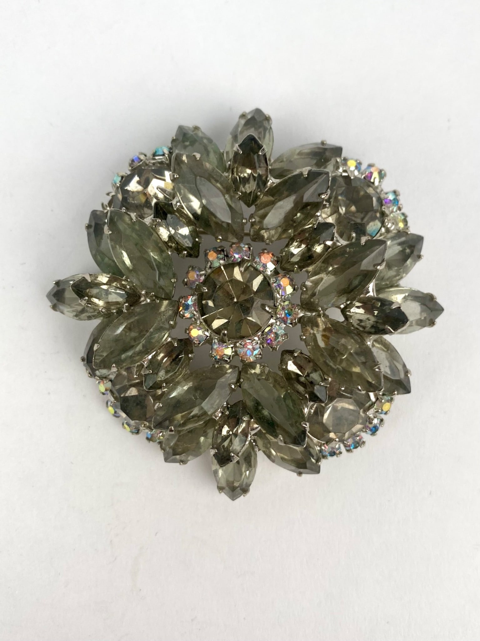 Mid Century Layered Rhinestone Brooch: Mid century brooch featuring layered tiers of smokey gray marquise rhinestones arranged in a floral burst motif. The center is encircled with iridescent aurora borealis rhinestones. The stones are set