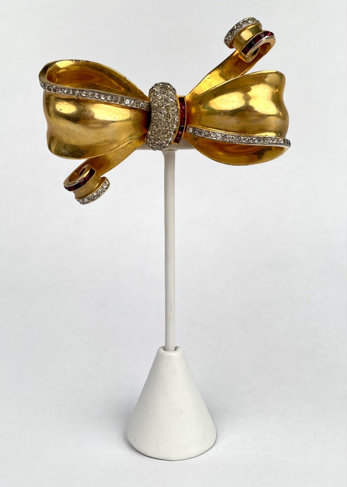1940s Gold Tone Bow Brooch with Rhinestones and Red Accents: 1940s costume jewelry brooch styled as a dimensional bow. Crafted in gold tone metal, the piece is richly adorned with sparkling clear rhinestones along the ribbon edges and center knot with red bague