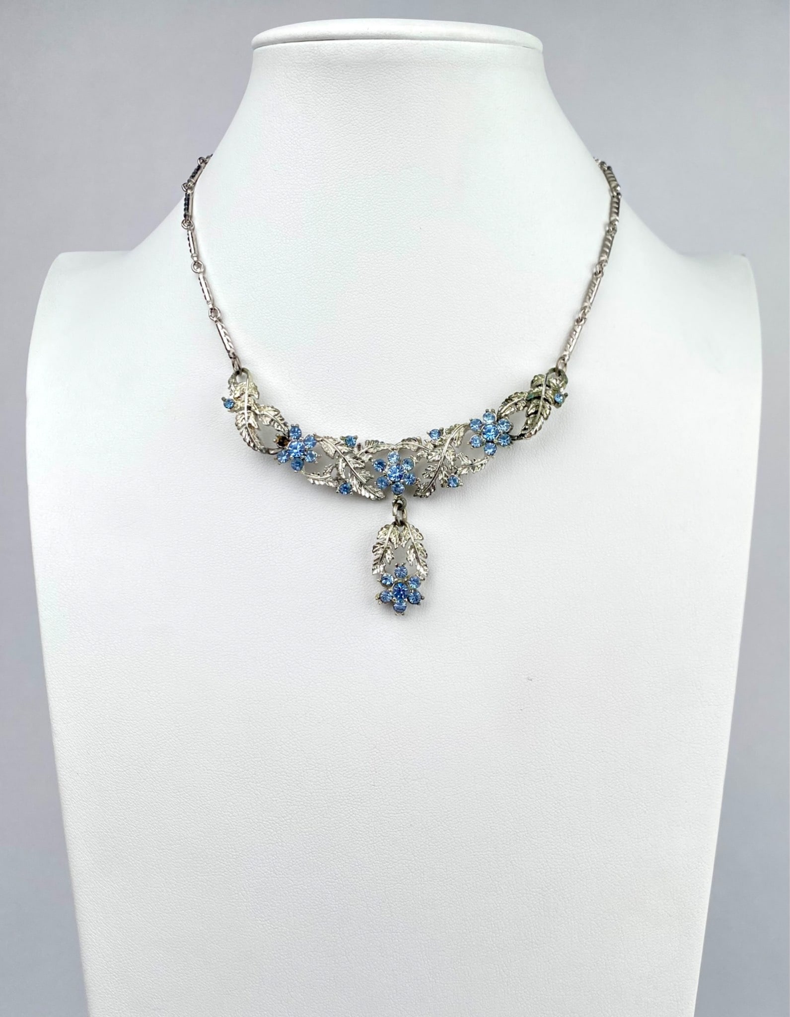 Mid Century Silver Tone Necklace with Blue Rhinestone Floral Motif: Silver tone necklace featuring an intricate floral motif adorned with blue rhinestones. The design includes detailed leaf patterns and a center pendant with matching blue rhinestones. The chain featur