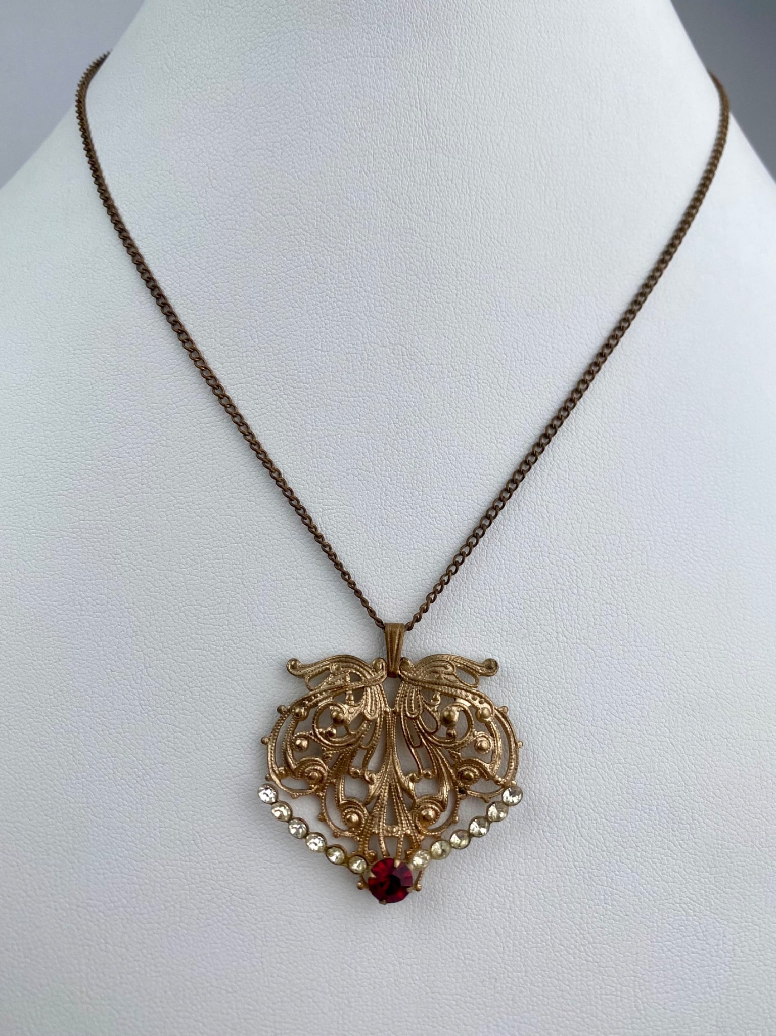 Victorian Revival Filigree Pendant Necklace with Red and Clear Rhinestones: Detailed Victorian Revival pendant necklace featuring an ornate gold tone filigree design. The centerpiece is adorned with row of clear rhinestones and a single red rhinestone at the base. The intrica